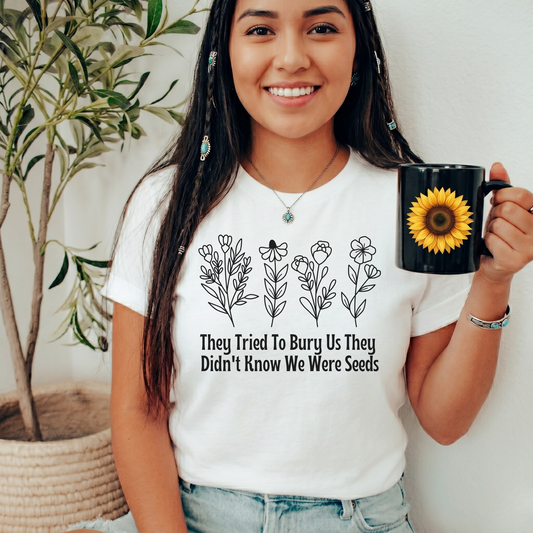 A woman wearing a white graphic t-shirt with black line art of flowers and the text 'They Tried To Bury Us They Didn't Know We Were Seeds', holding a black mug with a yellow sunflower print. She is standing indoors next to a potted plant.
