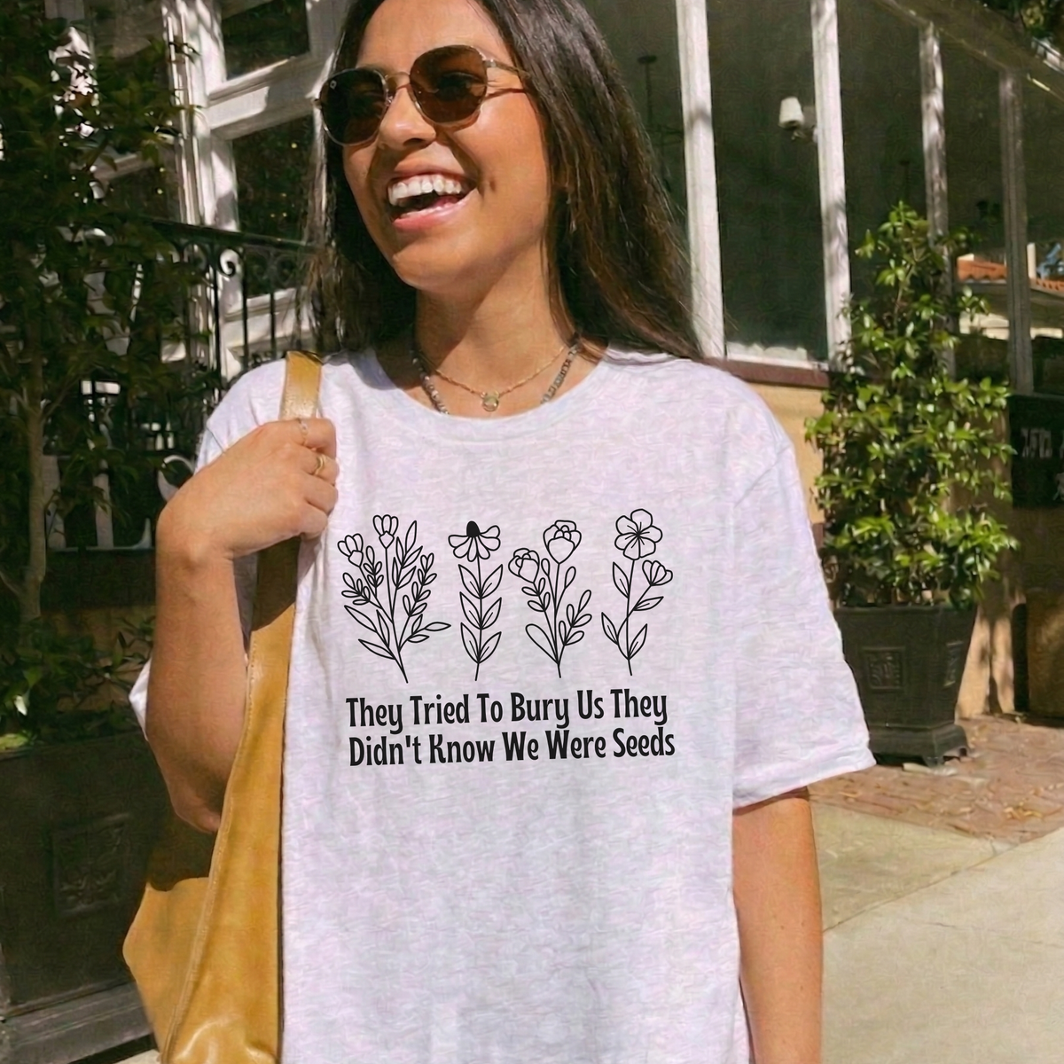 A woman wearing a light heather gray graphic t-shirt with a black floral design and the text 'They Tried To Bury Us They Didn't Know We Were Seeds'. She is outdoors on a sunny day, holding a tan shoulder bag.