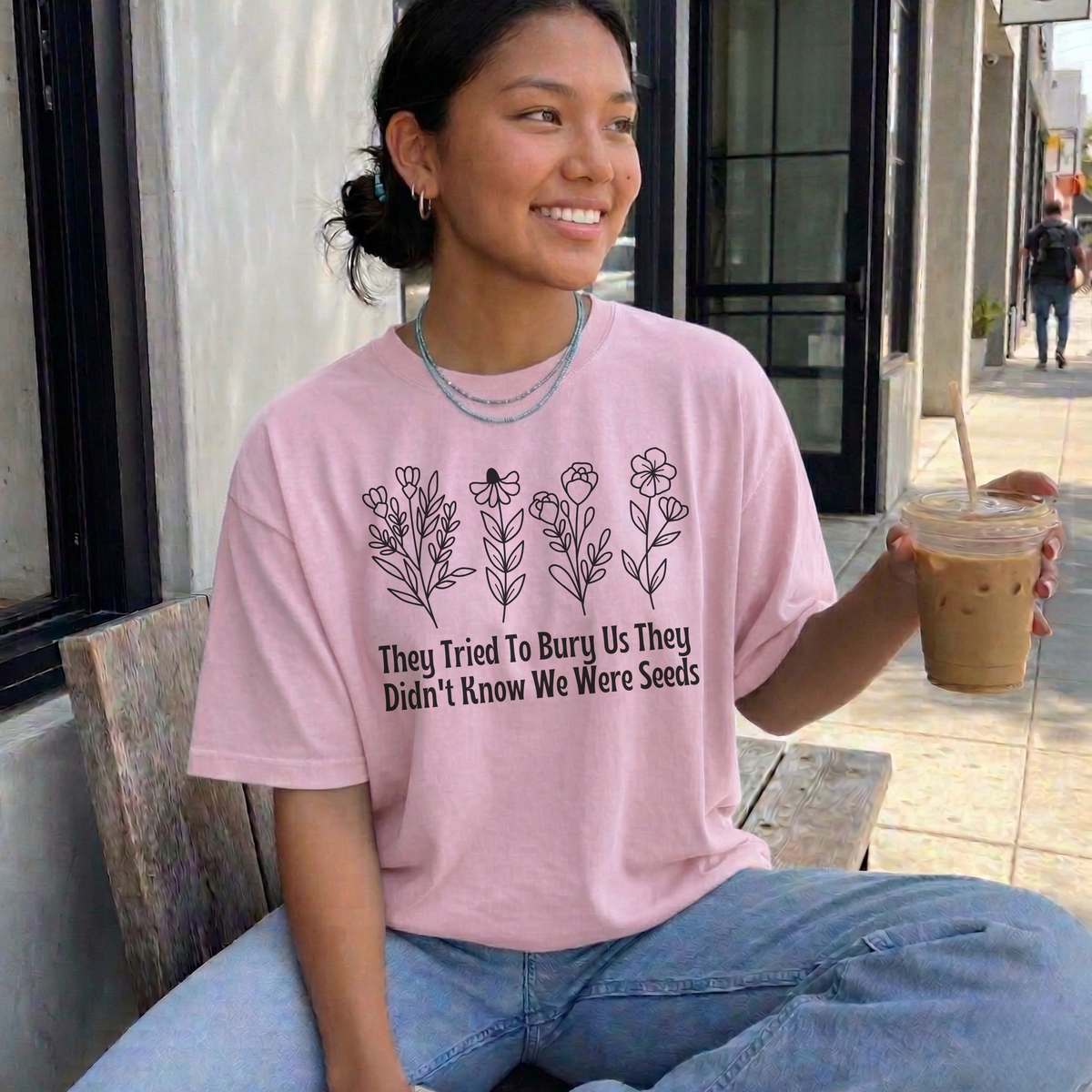 A woman sitting on a bench outdoors wearing a light pink graphic t-shirt with black text and floral illustrations. She is holding an iced coffee and wearing light blue jeans.