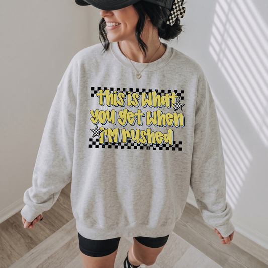 A woman wearing an oversized light gray sweatshirt with a graphic that reads 'this is what you get when i'm rushed' in yellow text over a black-and-white checkerboard background. She is also wearing a black cap and black shorts in a brightly lit indoor room.