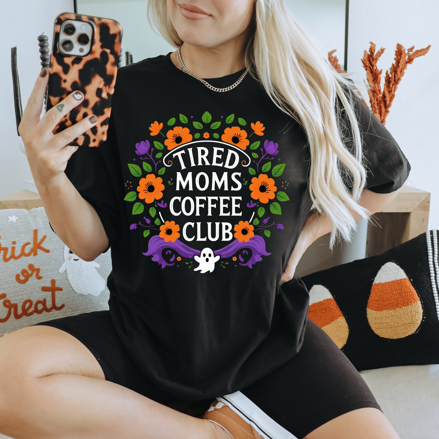 A woman takes a mirror selfie wearing a black graphic t-shirt with a colorful Halloween-themed design that reads 'TIRED MOMS COFFEE CLUB' with orange flowers, purple ghosts, and a small white ghost. She is wearing black biker shorts and holding a leopard-print phone case.