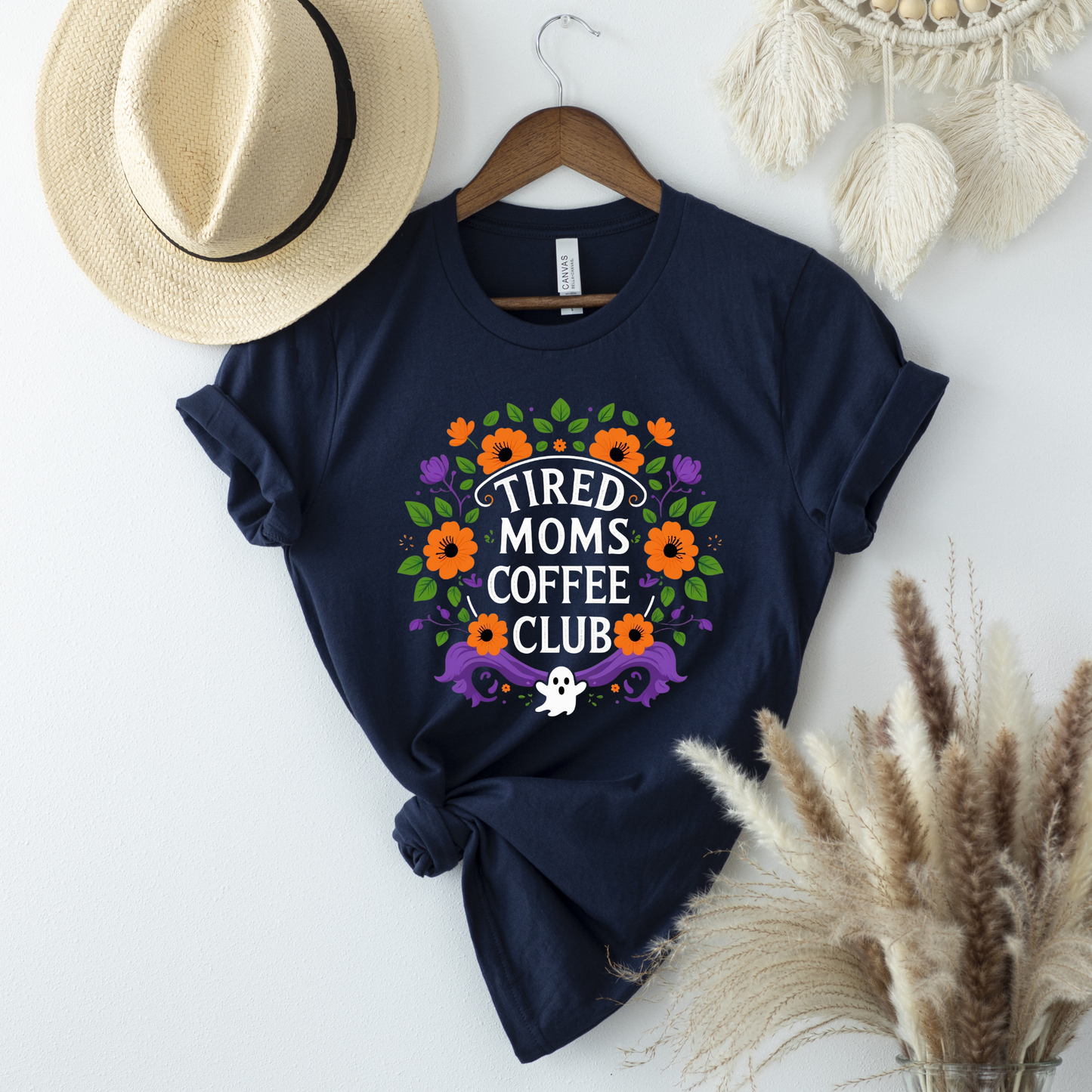 Navy blue t-shirt with floral design and text on a white surface with decorative items. tired moms coffee club, coffee mom shirt, funny mom tee, mom life shirt, unisex graphic tee, floral mom shirt, gift for mom, coffee lover shirt, casual mom wear, funny graphic shirt, everyday mom shirt, tired mom apparel, mom humor shirt, colorful floral tee, coffee club shirt