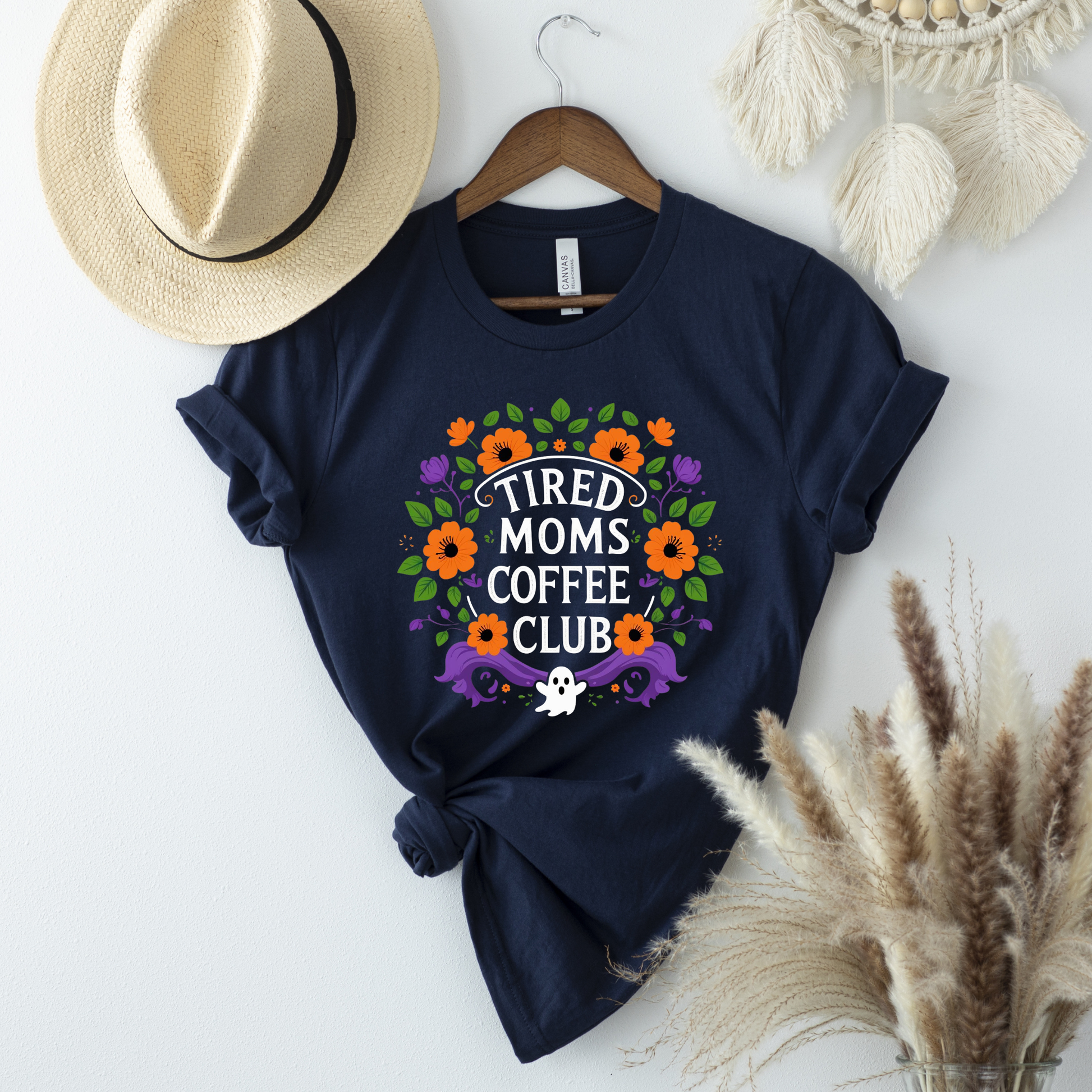Navy blue t-shirt with floral design and text on a white surface with decorative items. tired moms coffee club, coffee mom shirt, funny mom tee, mom life shirt, unisex graphic tee, floral mom shirt, gift for mom, coffee lover shirt, casual mom wear, funny graphic shirt, everyday mom shirt, tired mom apparel, mom humor shirt, colorful floral tee, coffee club shirt
