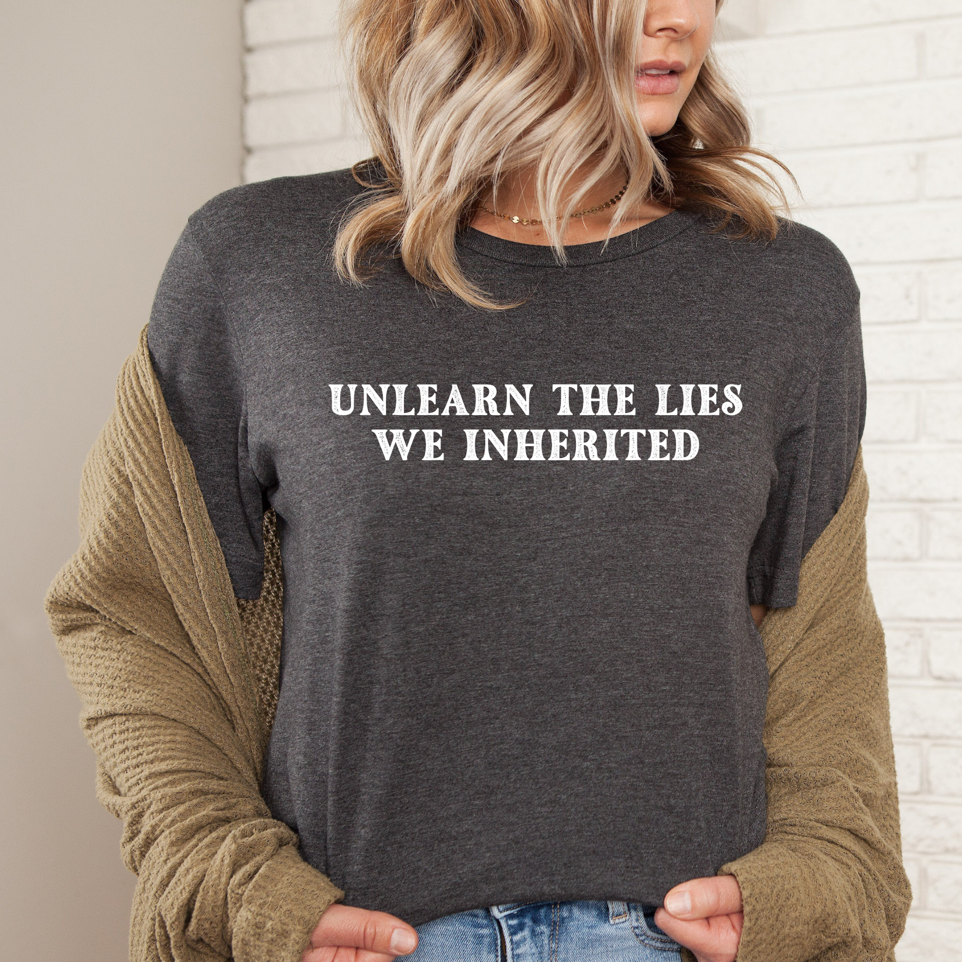 A woman wearing a dark heather gray long-sleeve t-shirt with the text 'UNLEARN THE LIES WE INHERITED' printed in white capital letters across the chest, layered with a tan knit cardigan. She is standing against a white brick wall background.
