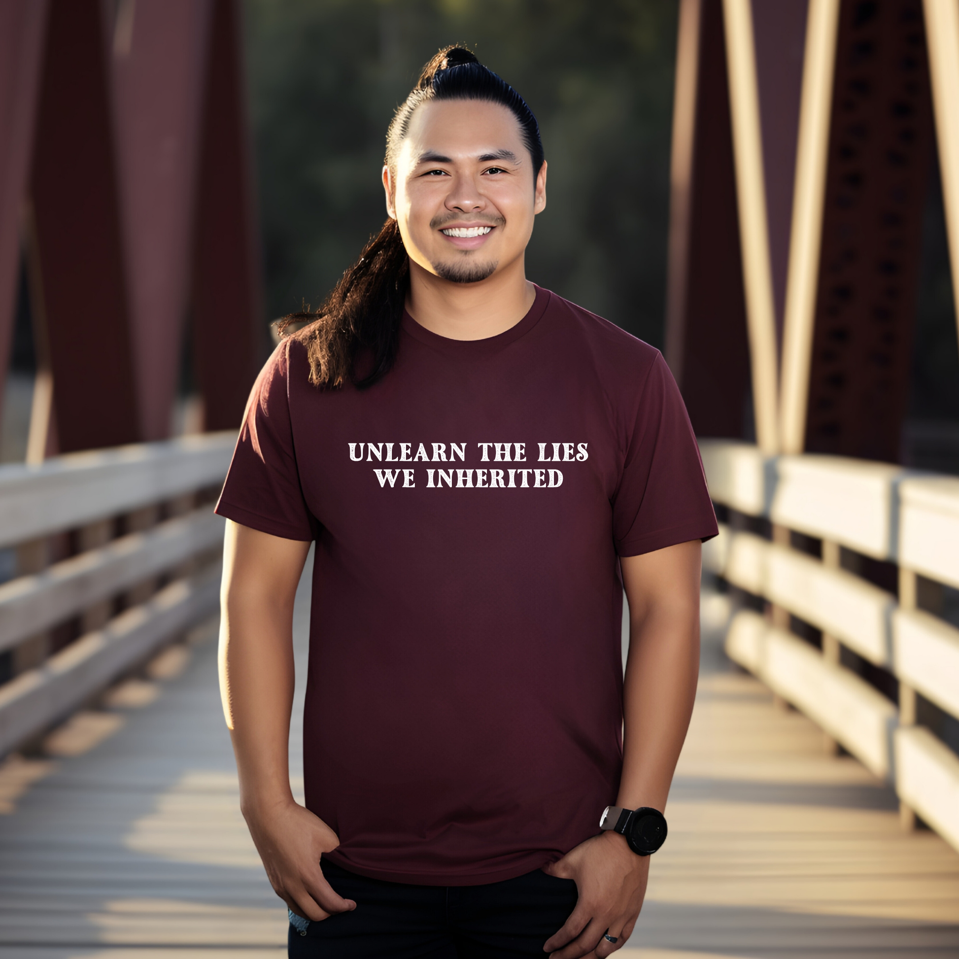 A man standing on a wooden bridge wearing a maroon t-shirt with the white printed text 'UNLEARN THE LIES WE INHERITED'. unlearn the lies we inherited, truth and reconciliation shirt, indigenous shirt, social justice shirt, decolonize shirt, history matters shirt, educational shirt, activist shirt, statement tee, indigenous ally shirt, minimalist graphic tee, teacher shirt, advocacy apparel