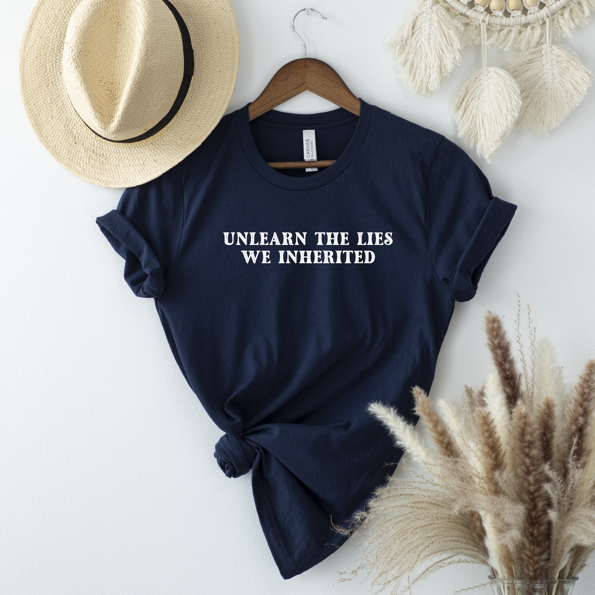 A navy blue short-sleeve t-shirt with white text reading 'UNLEARN THE LIES WE INHERITED' is displayed on a wooden hanger against a white wall, styled with a straw hat, a dreamcatcher, and dried pampas grass.