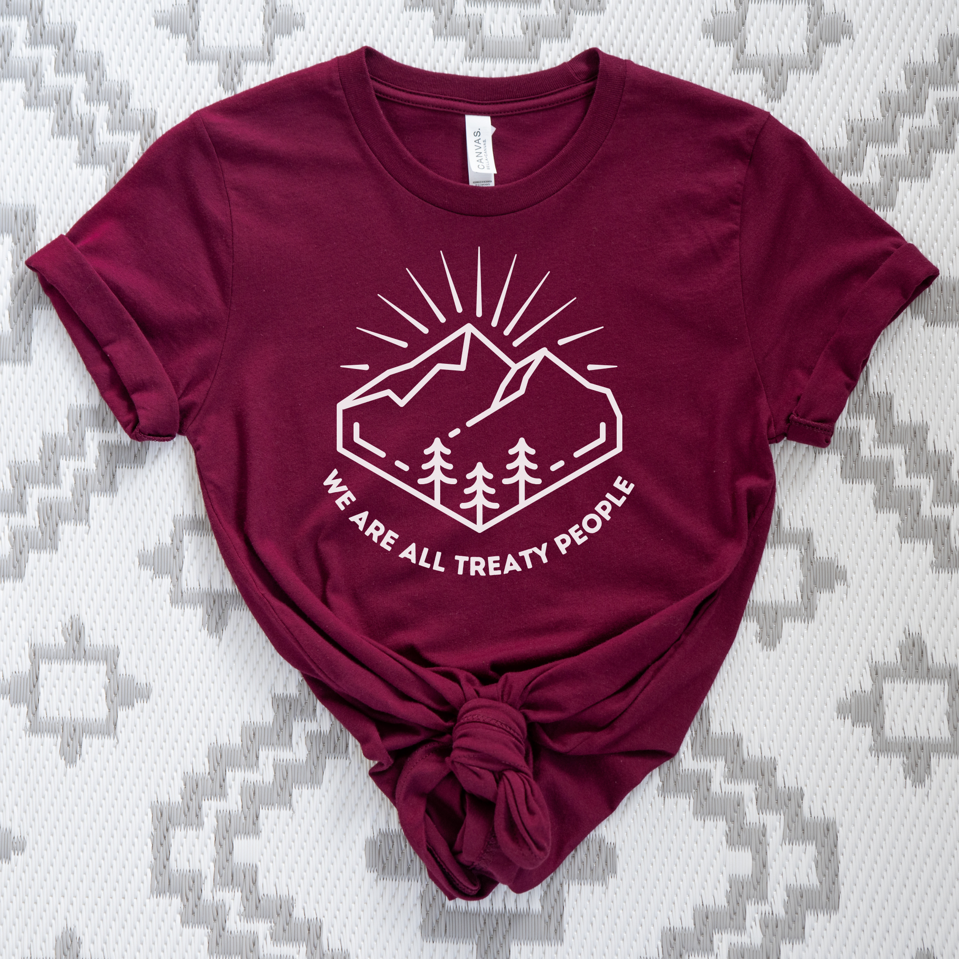 Maroon t-shirt with white graphic and text on a patterned surface. We Are All Treaty People, treaty shirt, Indigenous treaty awareness, Indigenous t shirt, unisex graphic tee, reconciliation shirt, Indigenous rights, awareness tee, activist shirt, Native apparel, solidarity shirt, truth and reconciliation, statement tee, Indigenous owned brand, educational shirt