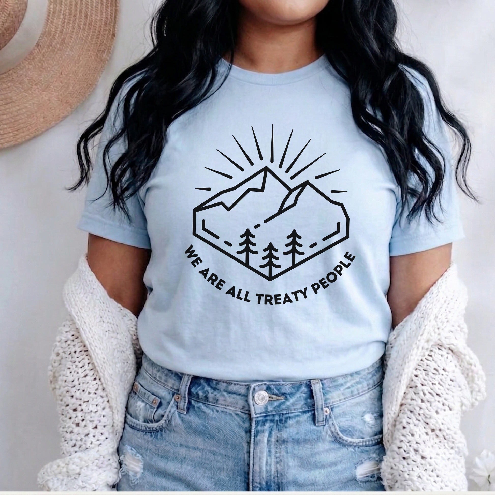 Person wearing a light blue t-shirt with a mountain and tree design and text. We Are All Treaty People, treaty shirt, Indigenous treaty awareness, Indigenous t shirt, unisex graphic tee, reconciliation shirt, Indigenous rights, awareness tee, activist shirt, Native apparel, solidarity shirt, truth and reconciliation, statement tee, Indigenous owned brand, educational shirt