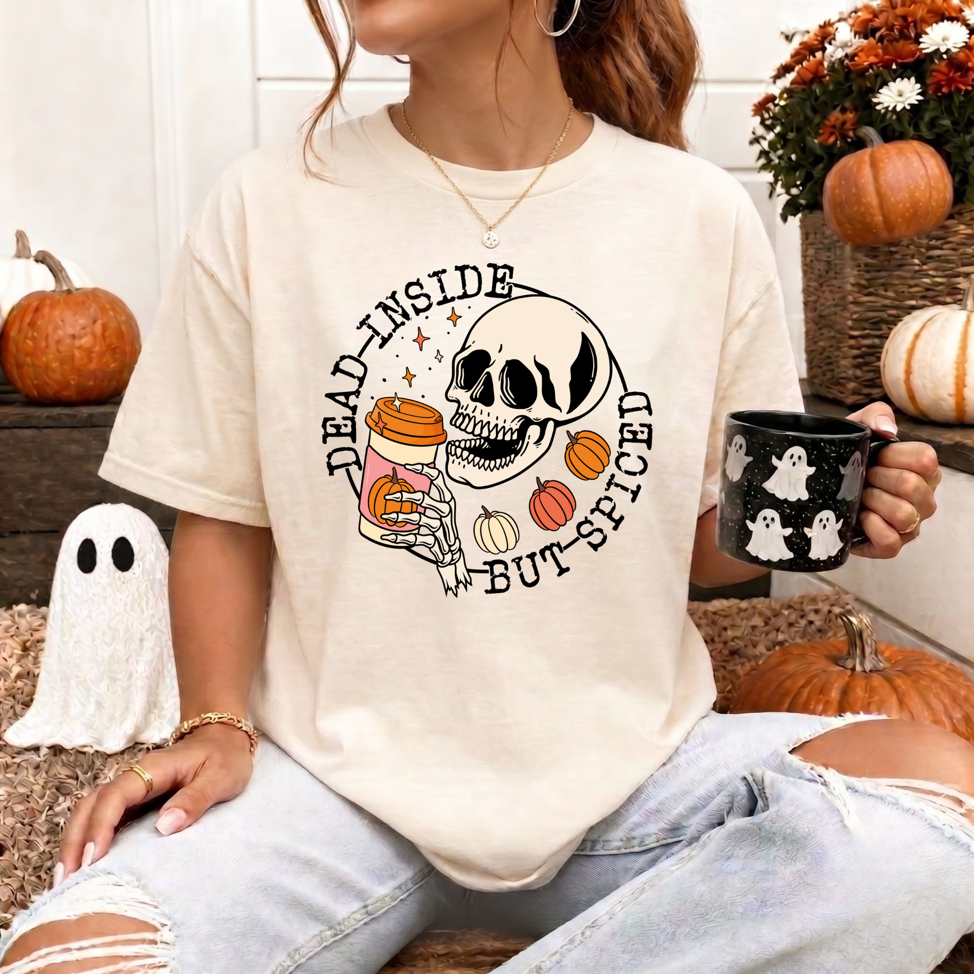 A woman wearing a cream-colored graphic t-shirt with a Halloween design featuring a skull, pumpkins, and the text 'DEAD INSIDE BUT SPICED'. She is holding a black mug with white ghosts, surrounded by autumn decorations like pumpkins and a ghost prop. dead inside but spiced, pumpkin spice shirt, skeleton coffee shirt, funny halloween shirt, spooky season shirt, fall coffee shirt, pumpkin patch outfit, halloween aesthetic, spooky coffee lover, skeleton tee, cozy fall shirt, autumn graphic tee, funny fall shir