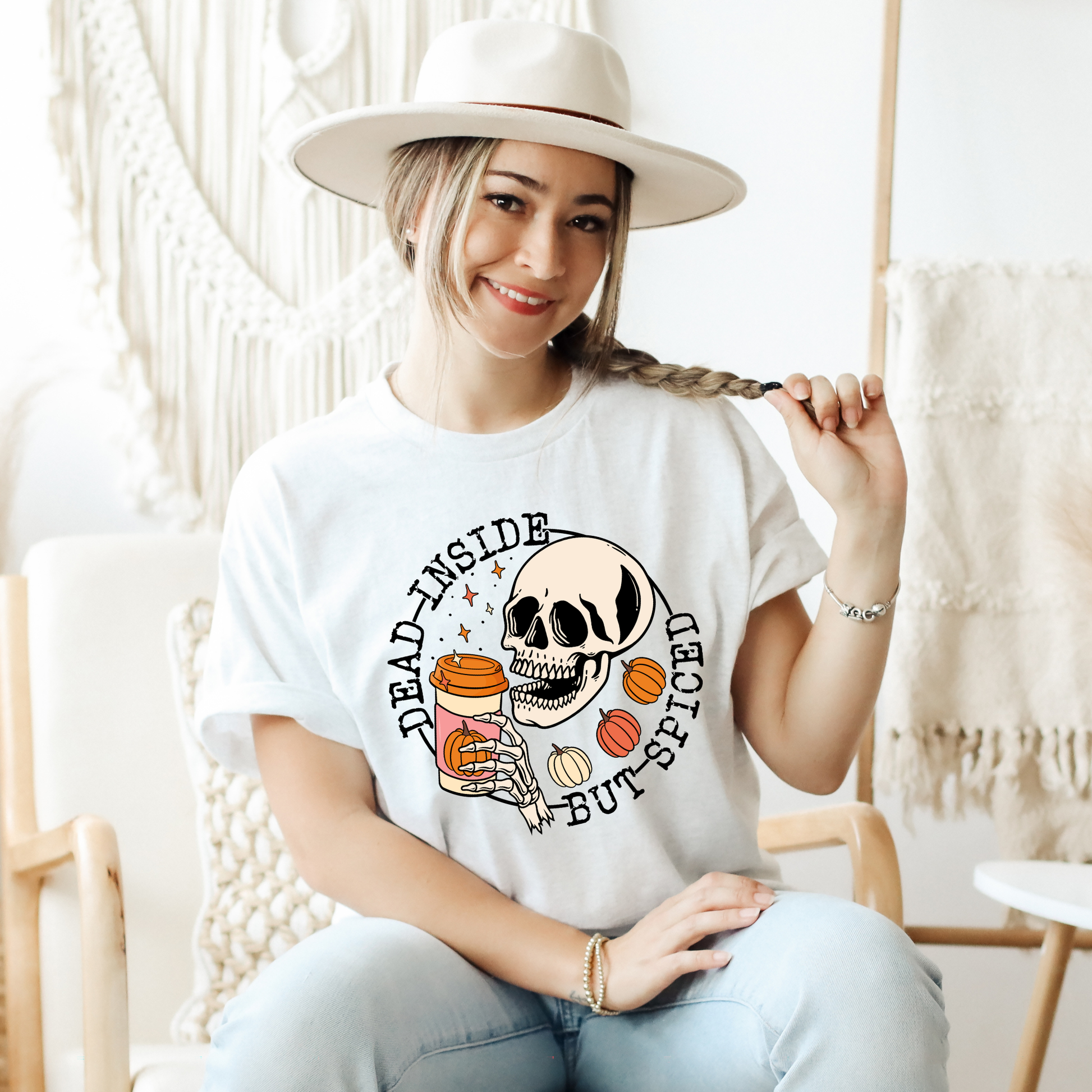 A woman wearing a white wide-brim hat and a white graphic t-shirt with a skull and food-themed print, sitting in a cozy, brightly lit room with macrame decor. dead inside but spiced, pumpkin spice shirt, skeleton coffee shirt, funny halloween shirt, spooky season shirt, fall coffee shirt, pumpkin patch outfit, halloween aesthetic, spooky coffee lover, skeleton tee, cozy fall shirt, autumn graphic tee, funny fall shirt