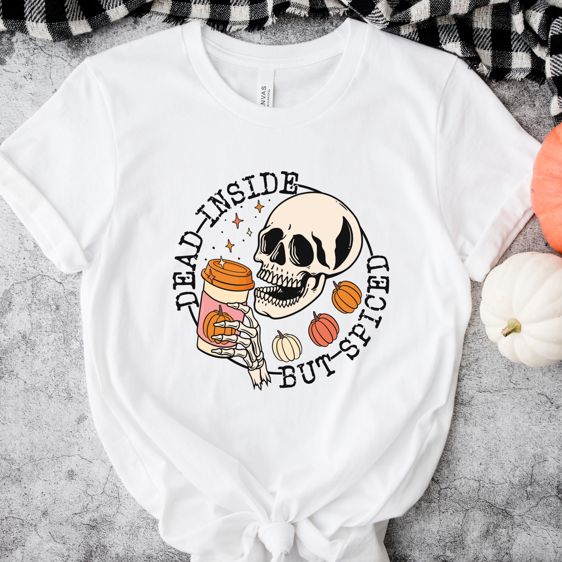 A white graphic t-shirt with a Halloween-themed design featuring a skull, pumpkins, and a coffee cup, styled flat-lay on a gray textured surface with a black-and-white plaid blanket and small pumpkins. dead inside but spiced, pumpkin spice shirt, skeleton coffee shirt, funny halloween shirt, spooky season shirt, fall coffee shirt, pumpkin patch outfit, halloween aesthetic, spooky coffee lover, skeleton tee, cozy fall shirt, autumn graphic tee, funny fall shirt