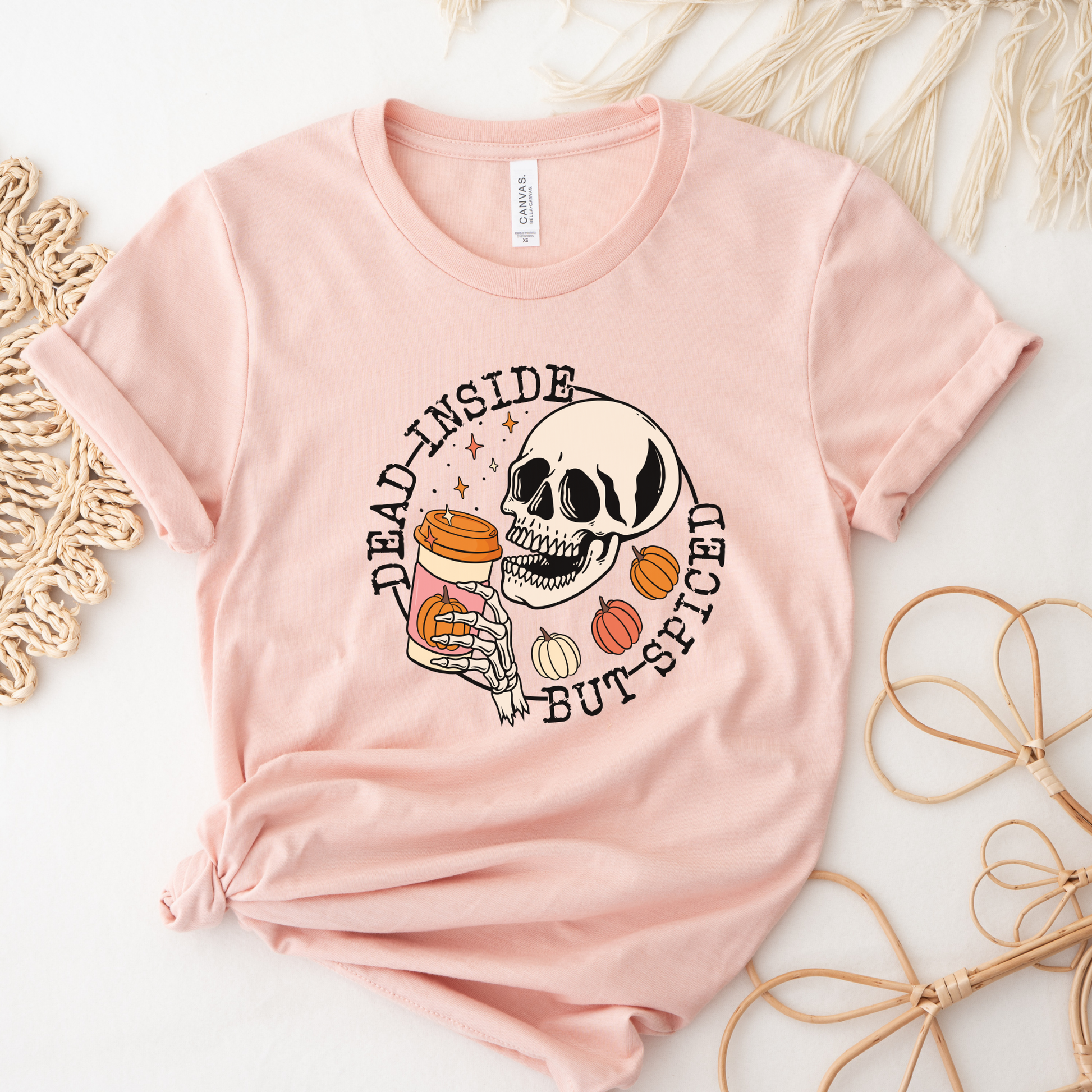A light pink short-sleeve t-shirt with a Halloween-themed graphic print featuring a skull, a jar of orange candies, and the text 'DEAD INSIDE BUT SPICED'. The shirt is styled in a flat-lay arrangement with neutral decorative props on a white background. 