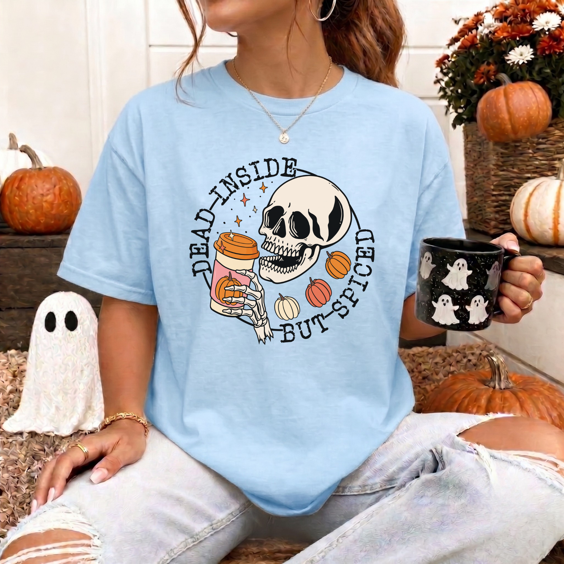 A woman wearing a light blue graphic t-shirt with a Halloween design featuring a skull, pumpkins, and the text 'DEAD INSIDE BUT SPICED'. She is holding a black mug with white ghost prints, sitting in a cozy, decorated autumn setting with pumpkins and a ghost decoration.