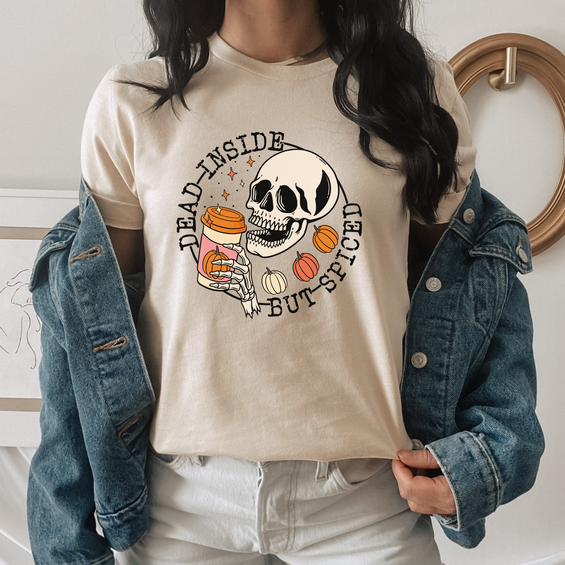 A person wearing a beige graphic T-shirt with a Halloween-themed skull and spice print, styled with an open blue denim jacket and white pants. dead inside but spiced, pumpkin spice shirt, skeleton coffee shirt, funny halloween shirt, spooky season shirt, fall coffee shirt, pumpkin patch outfit, halloween aesthetic, spooky coffee lover, skeleton tee, cozy fall shirt, autumn graphic tee, funny fall shirt