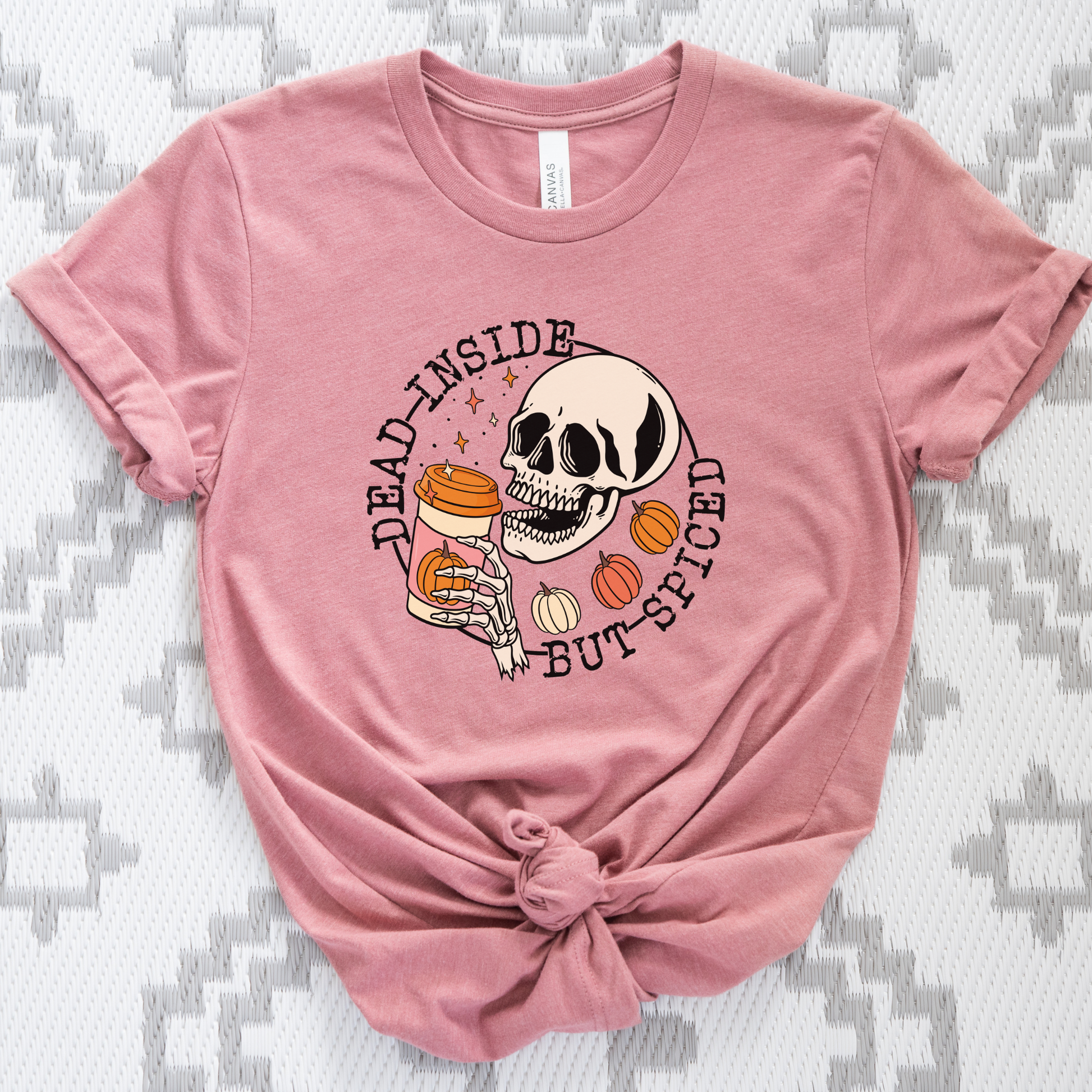 A dusty pink short-sleeve t-shirt with a graphic of a skull holding a jar of hot sauce, surrounded by the text 'DEAD INSIDE BUT SPICED', styled with a knot at the bottom hem on a gray and white patterned fabric background.