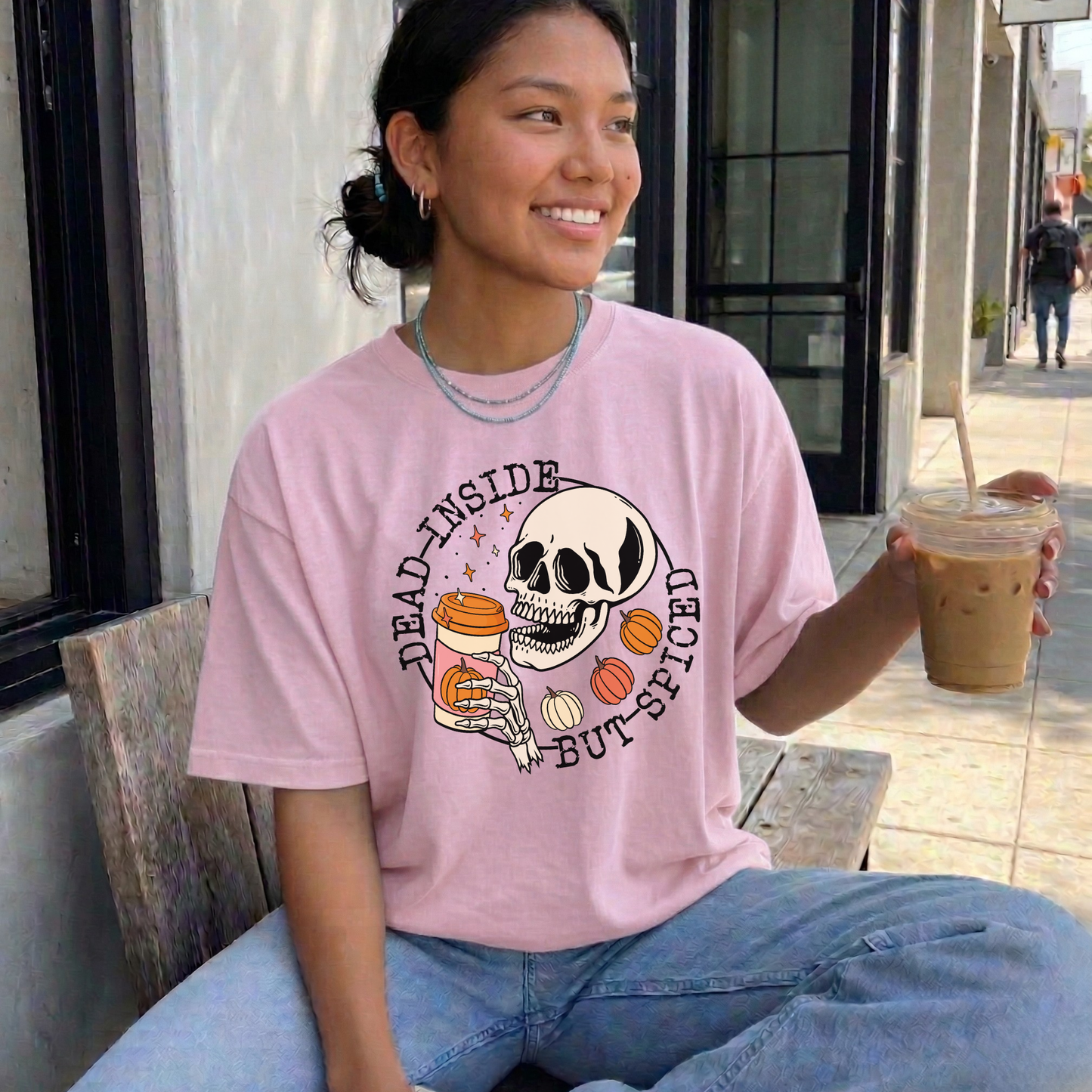 A woman sitting on a bench outdoors wearing a light pink graphic t-shirt with a skull and candy design and the text 'DRAG INSIDE BUT SPICED'. She is holding an iced coffee.