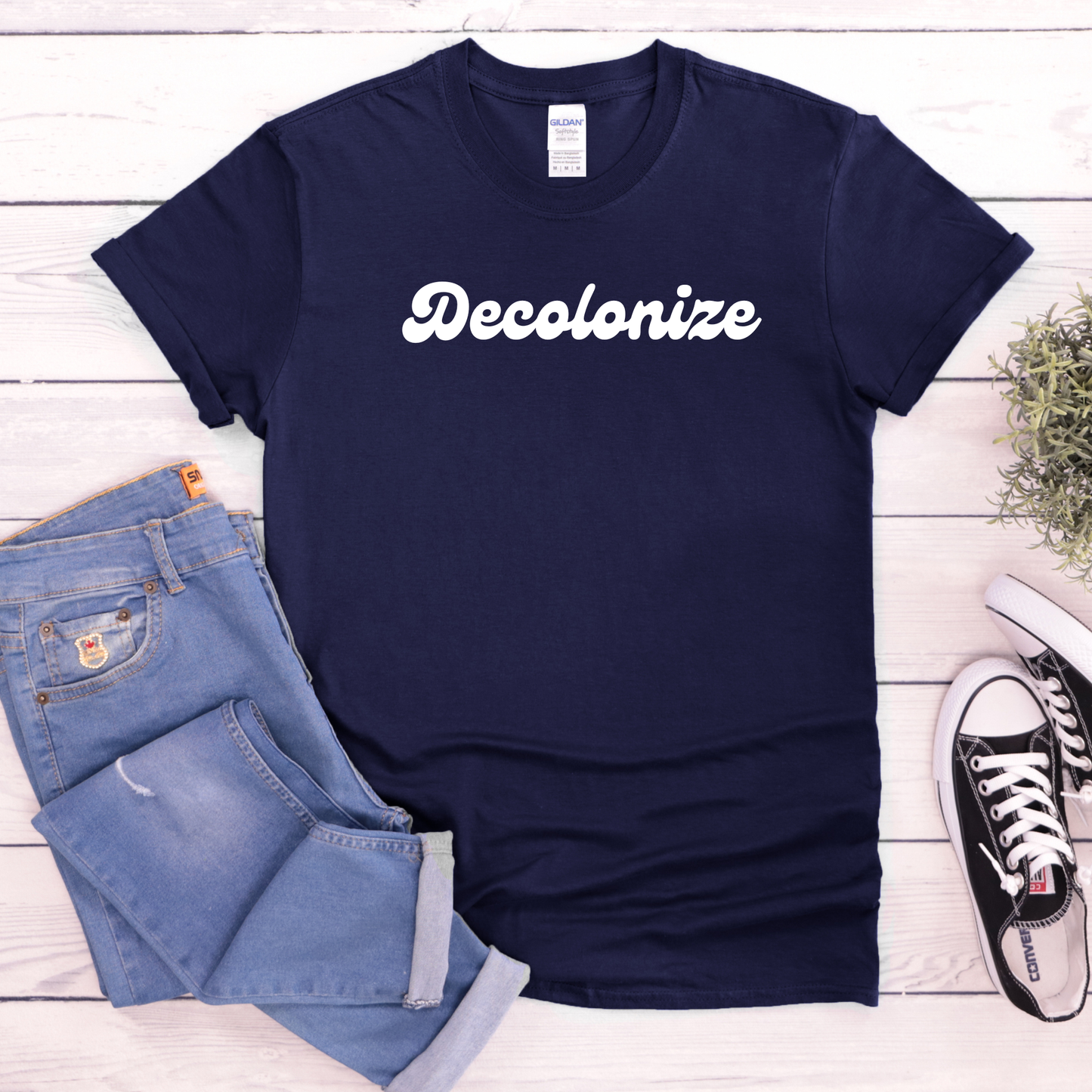 A navy blue t-shirt with the word 'Decolonize' printed in white script across the chest, styled in a flat lay with light blue jeans, black and white sneakers, and a small plant on a white wood plank background.