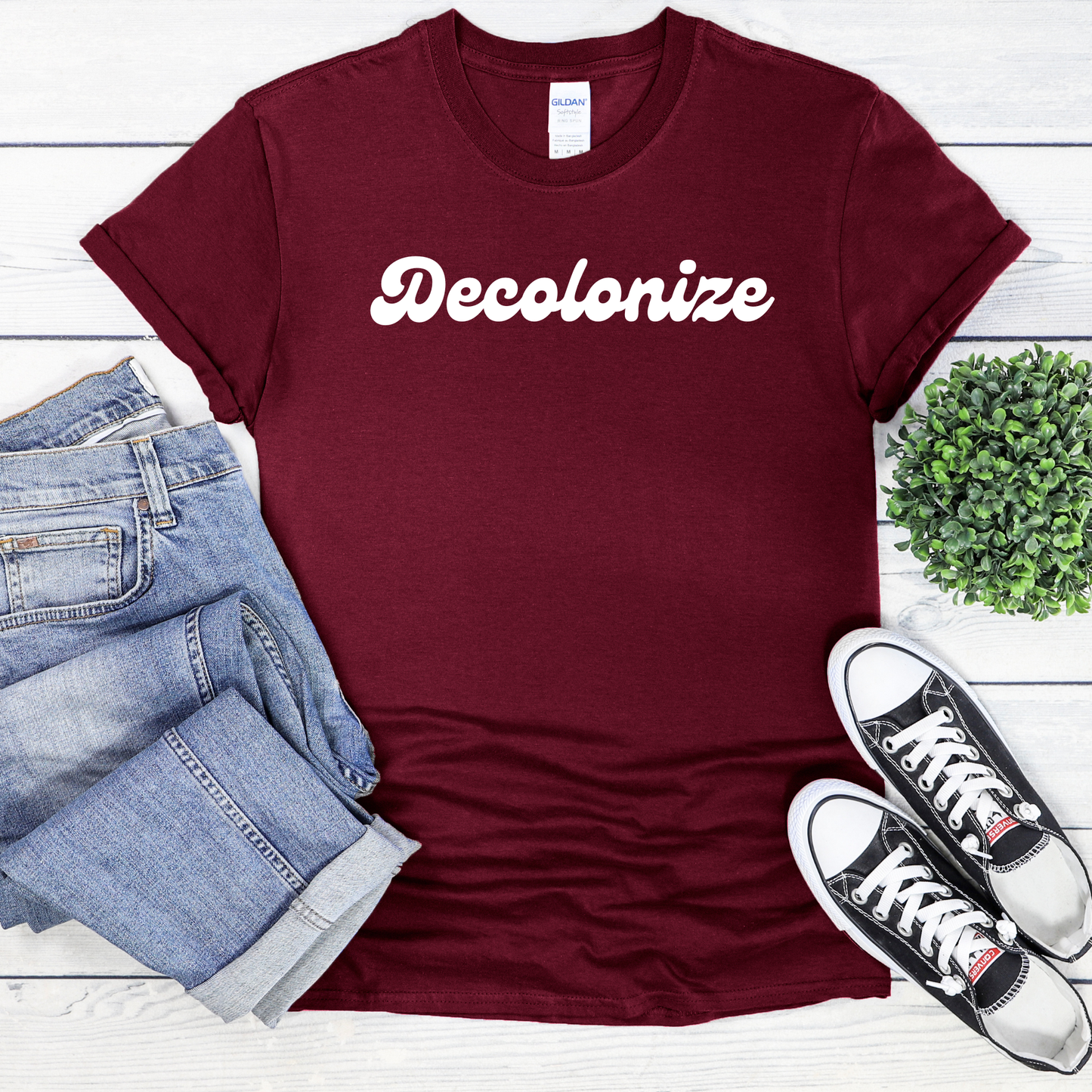 A maroon short-sleeve t-shirt with the word 'Decolonize' printed in white script across the chest, styled in a flat lay with light-wash jeans, black sneakers, and a small green plant on a white wood background.