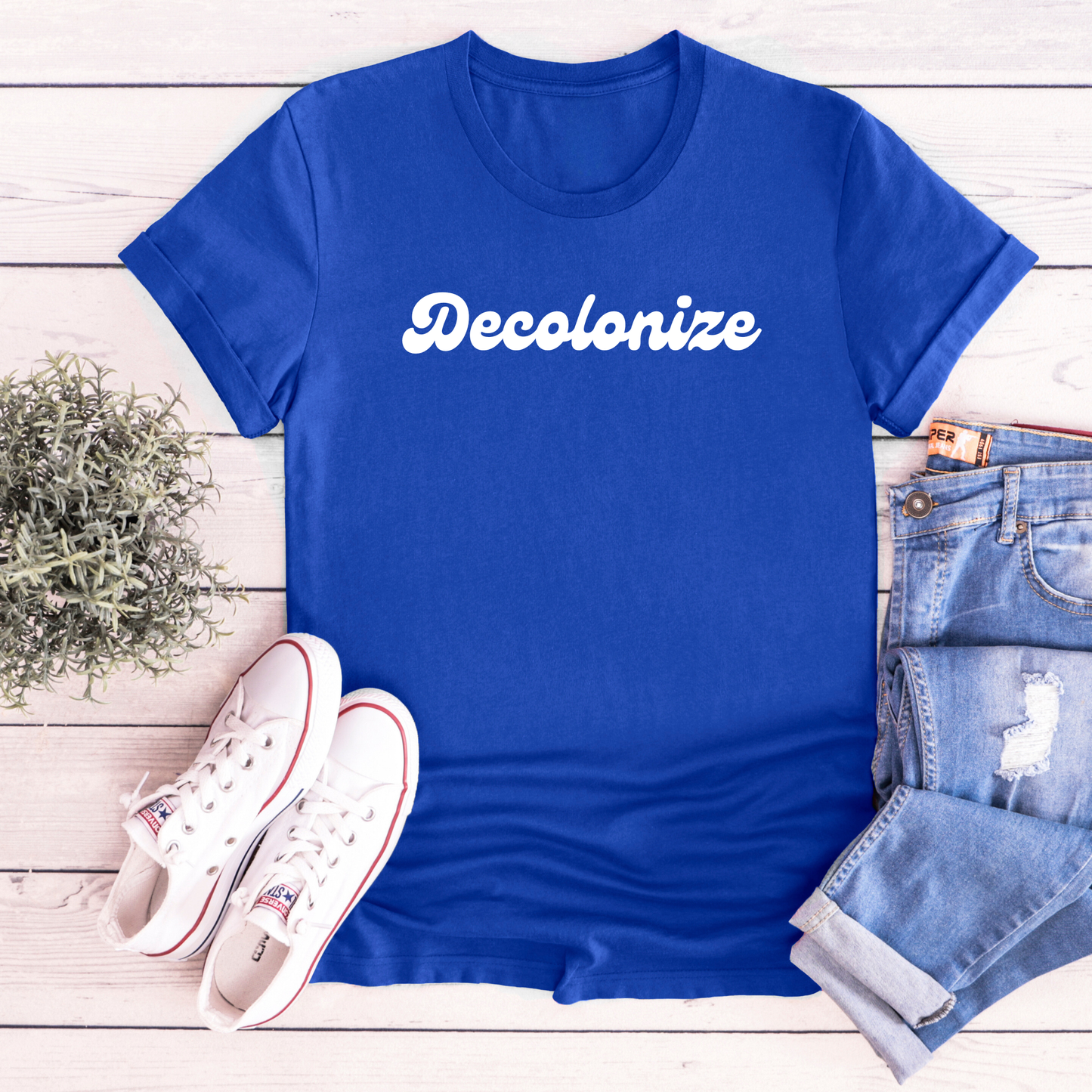 A royal blue short-sleeve t-shirt with the word 'Decolonize' printed in white script across the chest, styled in a flat lay on a white wood plank background with white sneakers, blue jeans, and a small green plant.