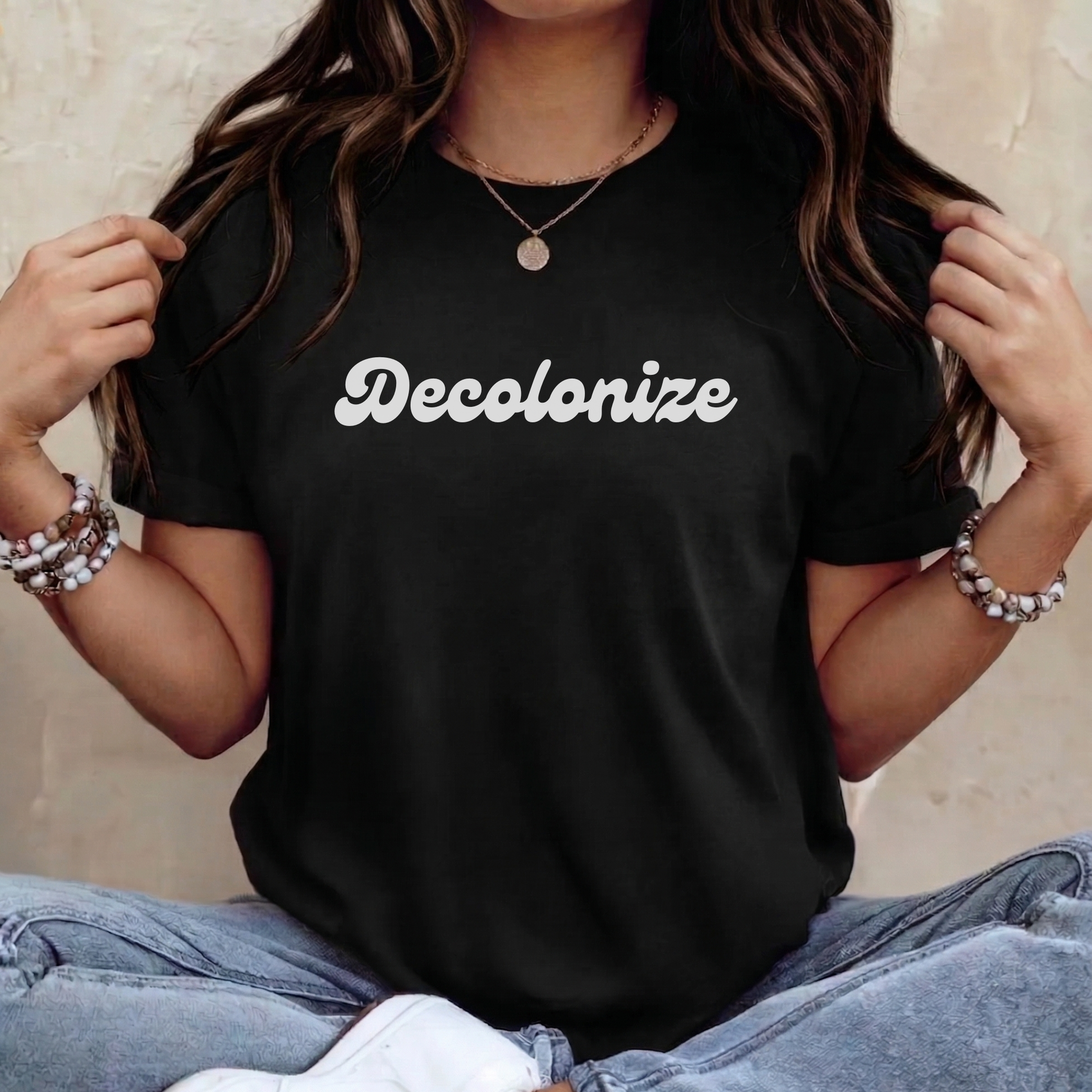 A woman wearing a black short-sleeve t-shirt with the word 'Decolonize' printed in white script across the chest, paired with light blue jeans and layered bracelets, seated against a neutral textured wall.
