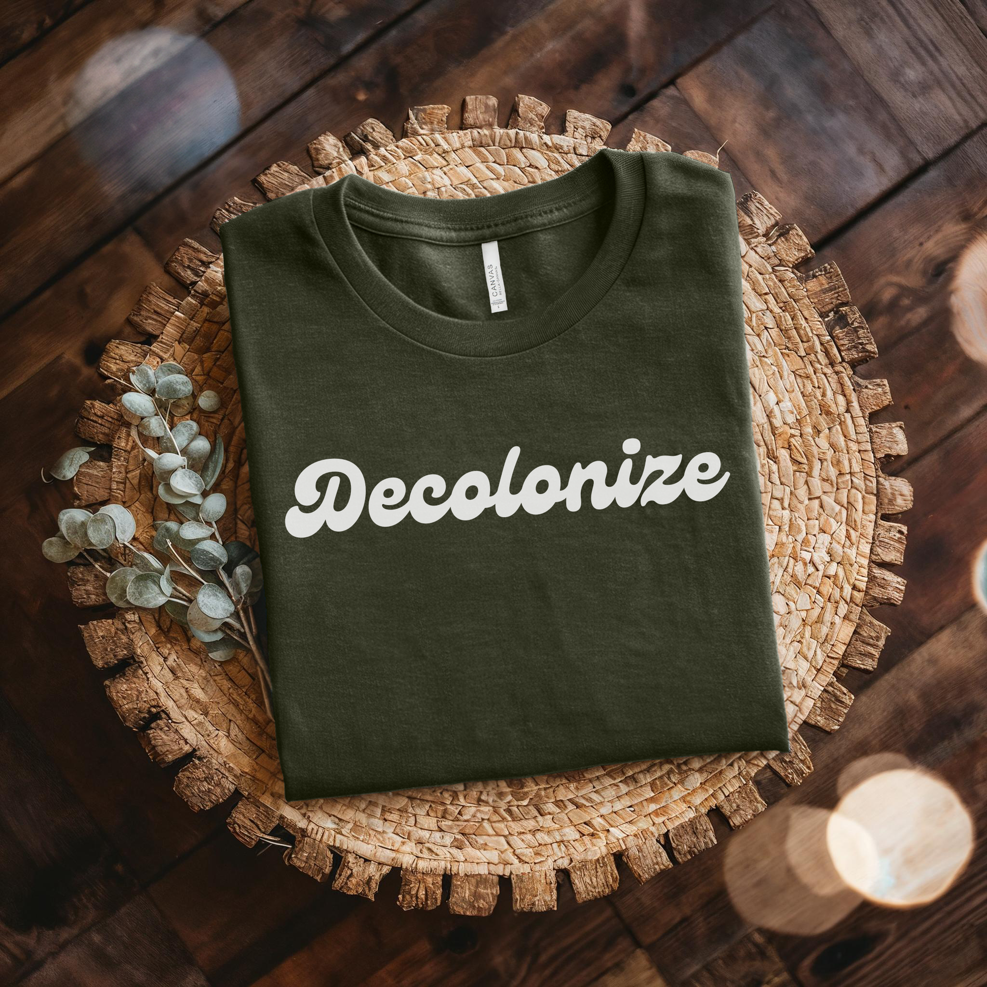 An olive green folded t-shirt with the word 'Decolonize' printed in white script across the chest, displayed on a round woven placemat over a dark wood background with decorative eucalyptus stems and soft bokeh lights.
