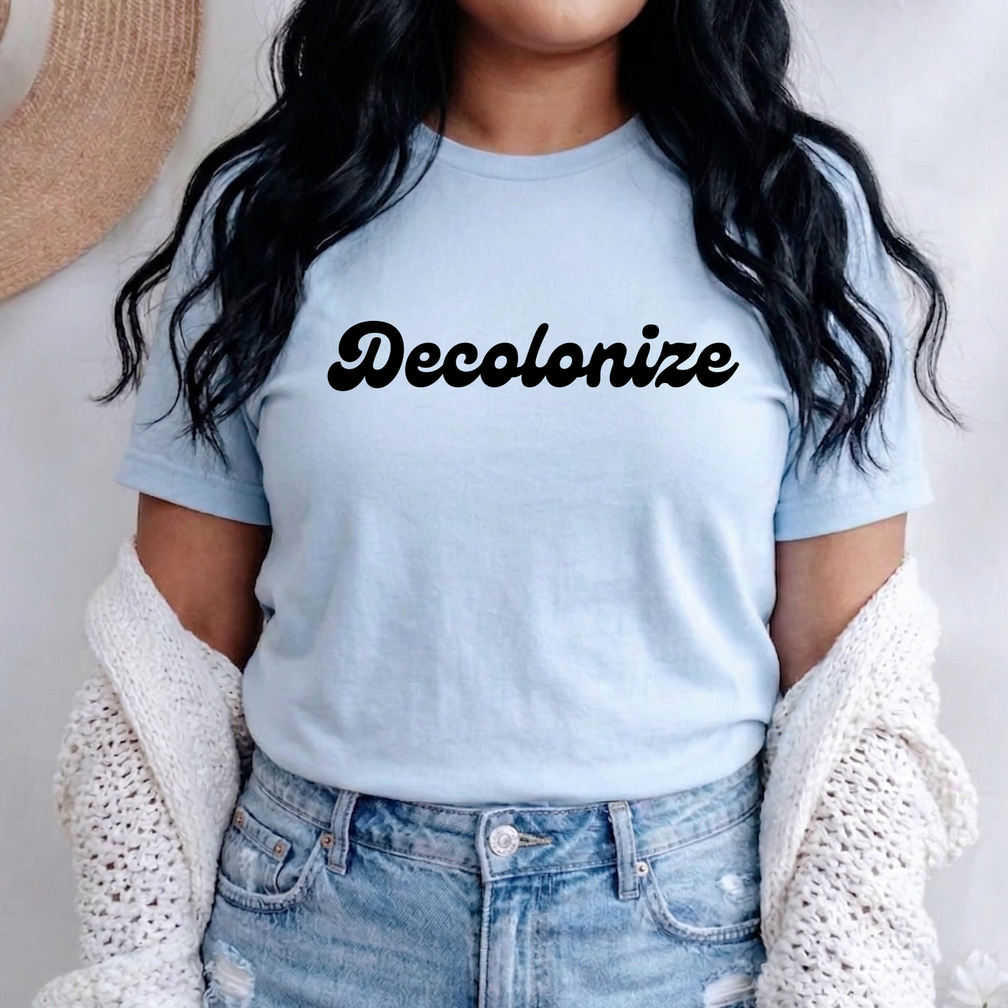 A woman wearing a light blue crewneck t-shirt with the word 'Decolonize' printed in black script across the chest, styled with a white knit cardigan and light-wash jeans.