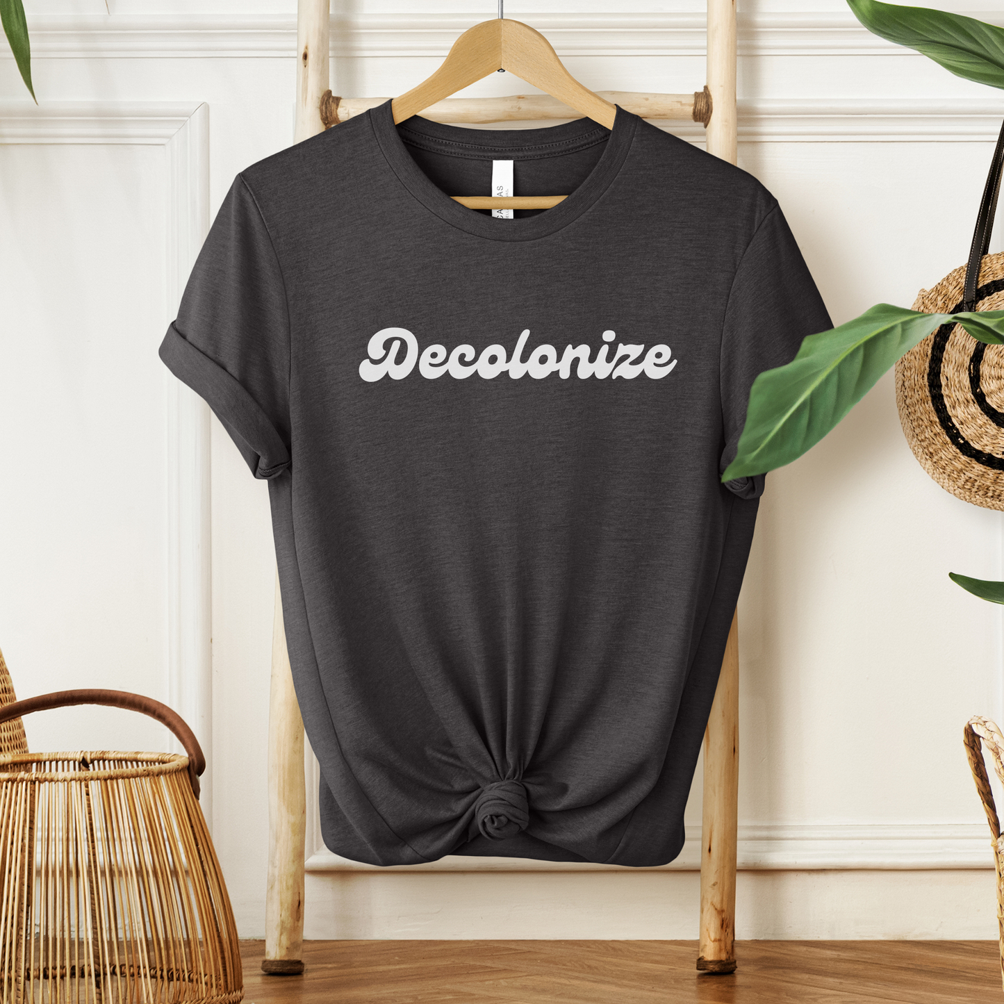 A dark charcoal gray t-shirt with the word 'Decolonize' printed in white script across the chest, styled on a wooden hanger with a knot at the bottom hem, set against a warm, boho-style background with plants and wicker.