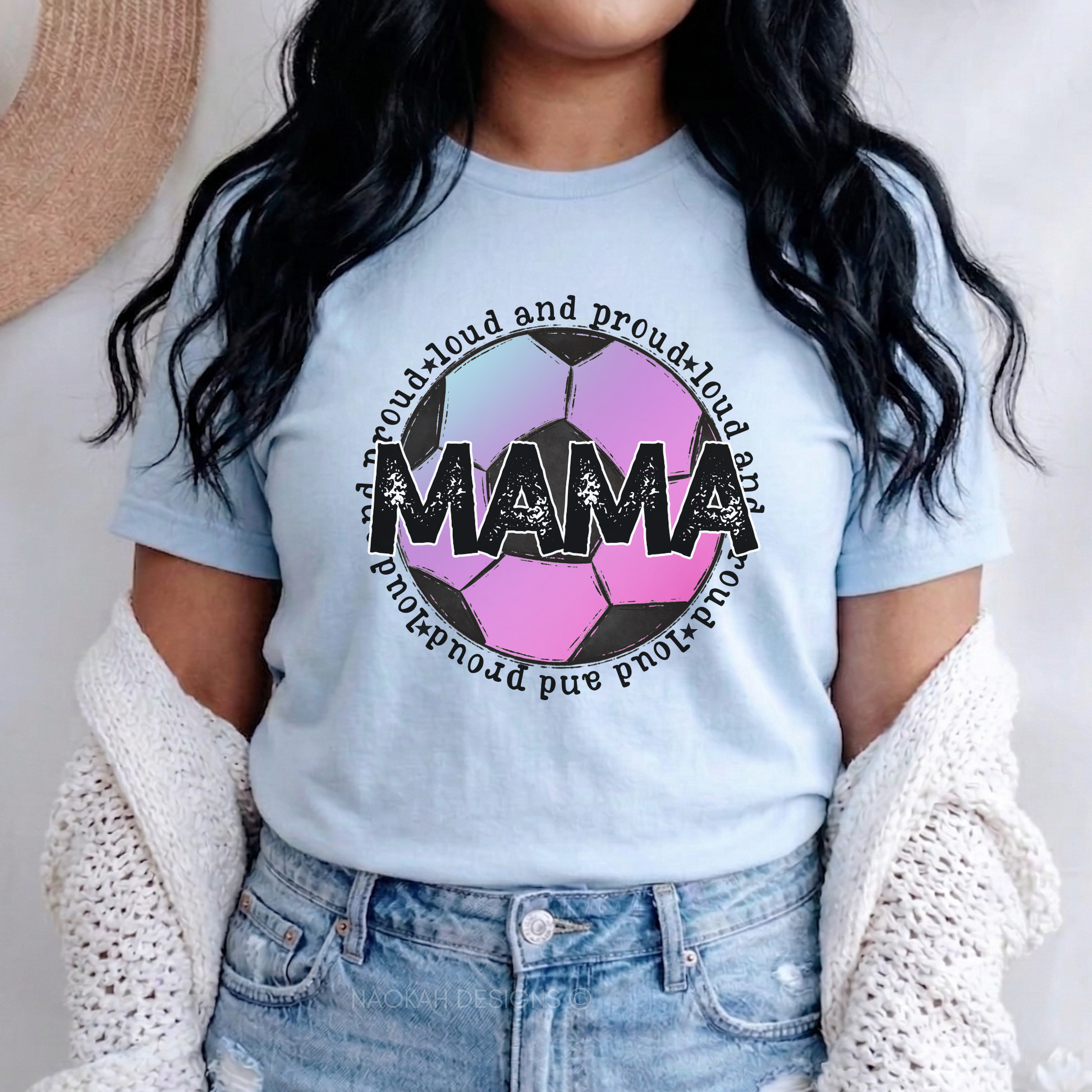 A woman wearing a light blue graphic t-shirt with a pink and black soccer ball design and the word 'MAMA' in the center, styled with a white knit cardigan and blue jeans.
