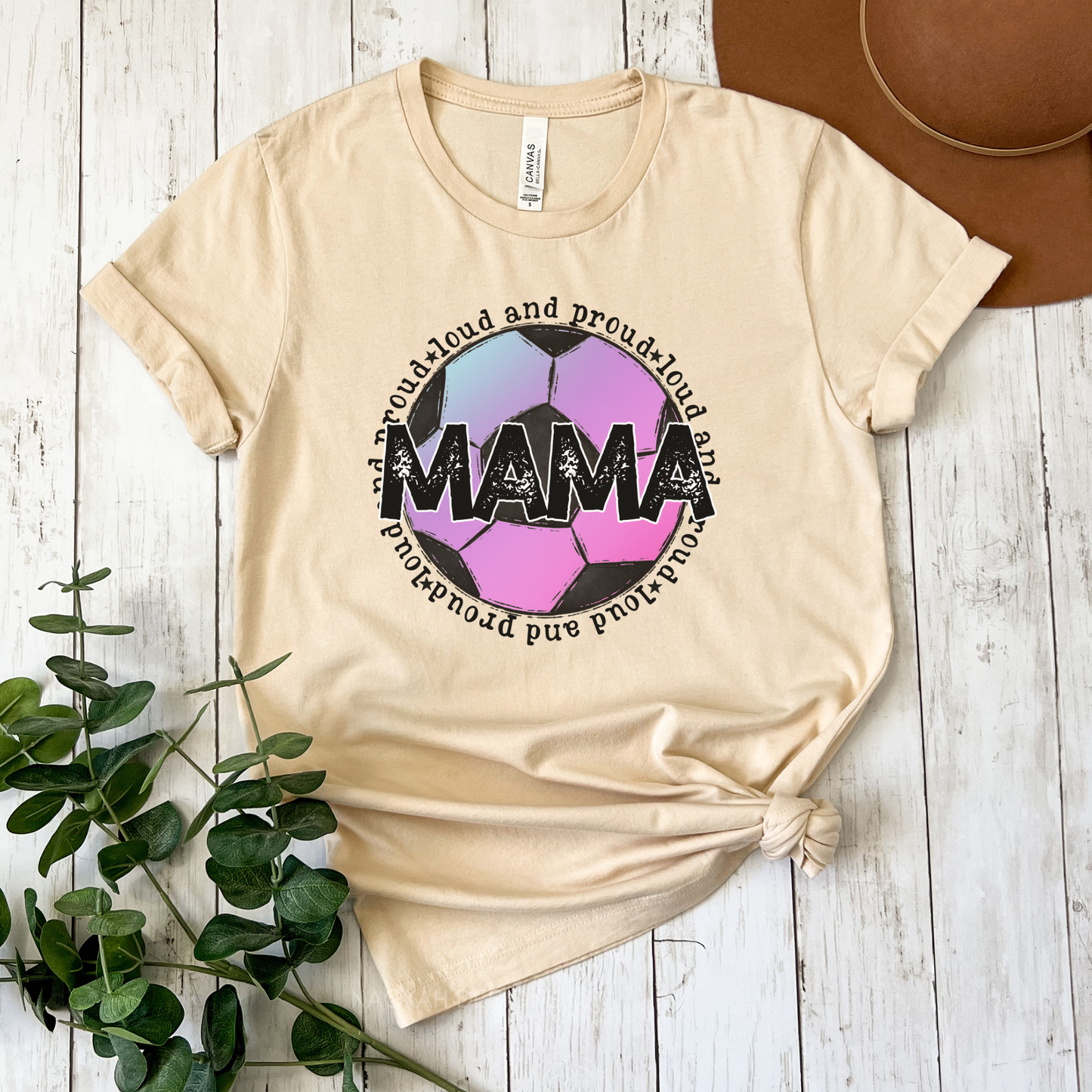 A beige graphic t-shirt featuring a soccer ball design with the word 'MAMA' and the phrase 'proud and proud of my mama' in black, styled in a flat-lay on a white wood background with a brown hat and green eucalyptus leaves.