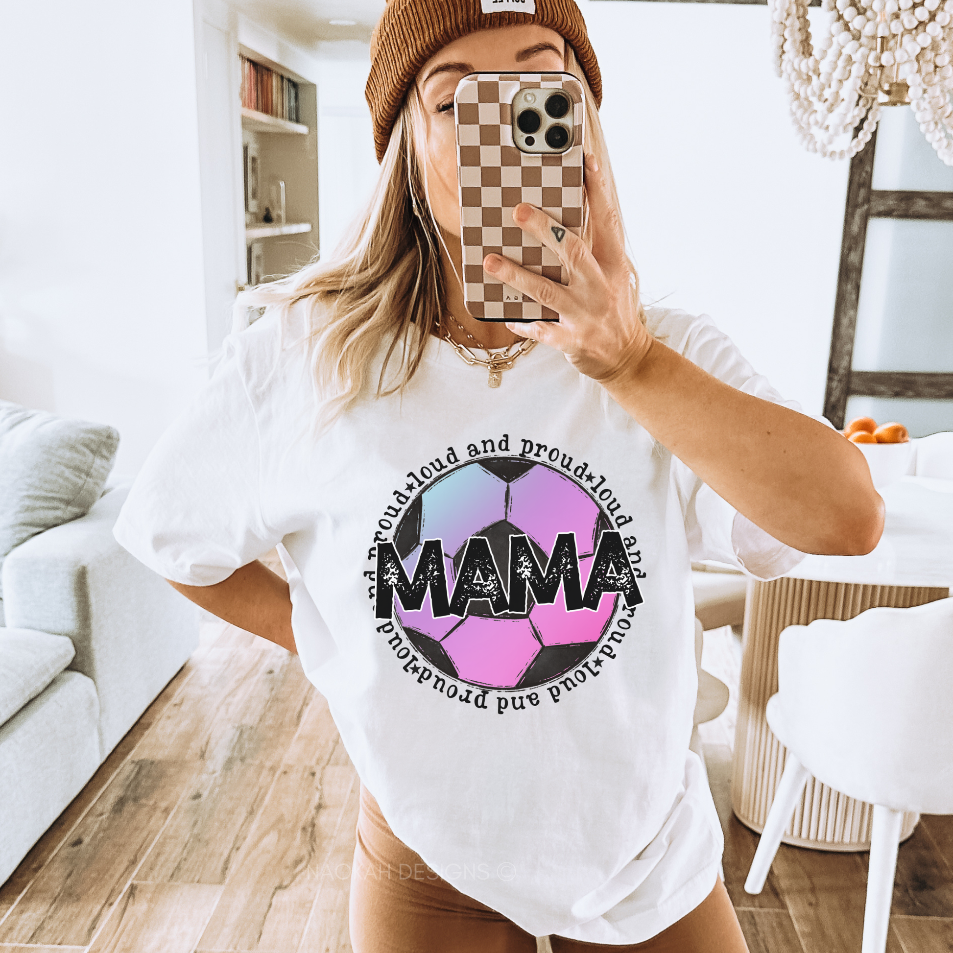 A woman takes a mirror selfie in a bright living room, wearing a white graphic t-shirt with a colorful soccer ball design and the word 'MAMA' printed on the front, paired with a brown beanie and tan pants. loud and proud soccer mom, soccer mom shirt, mama soccer tee, game day shirt, soccer mom gift, sports mom shirt, soccer season shirt, distressed mama shirt, soccer tournament outfit, athletic mom shirt, soccer fan apparel, soccer graphic tee, soccer mom apparel