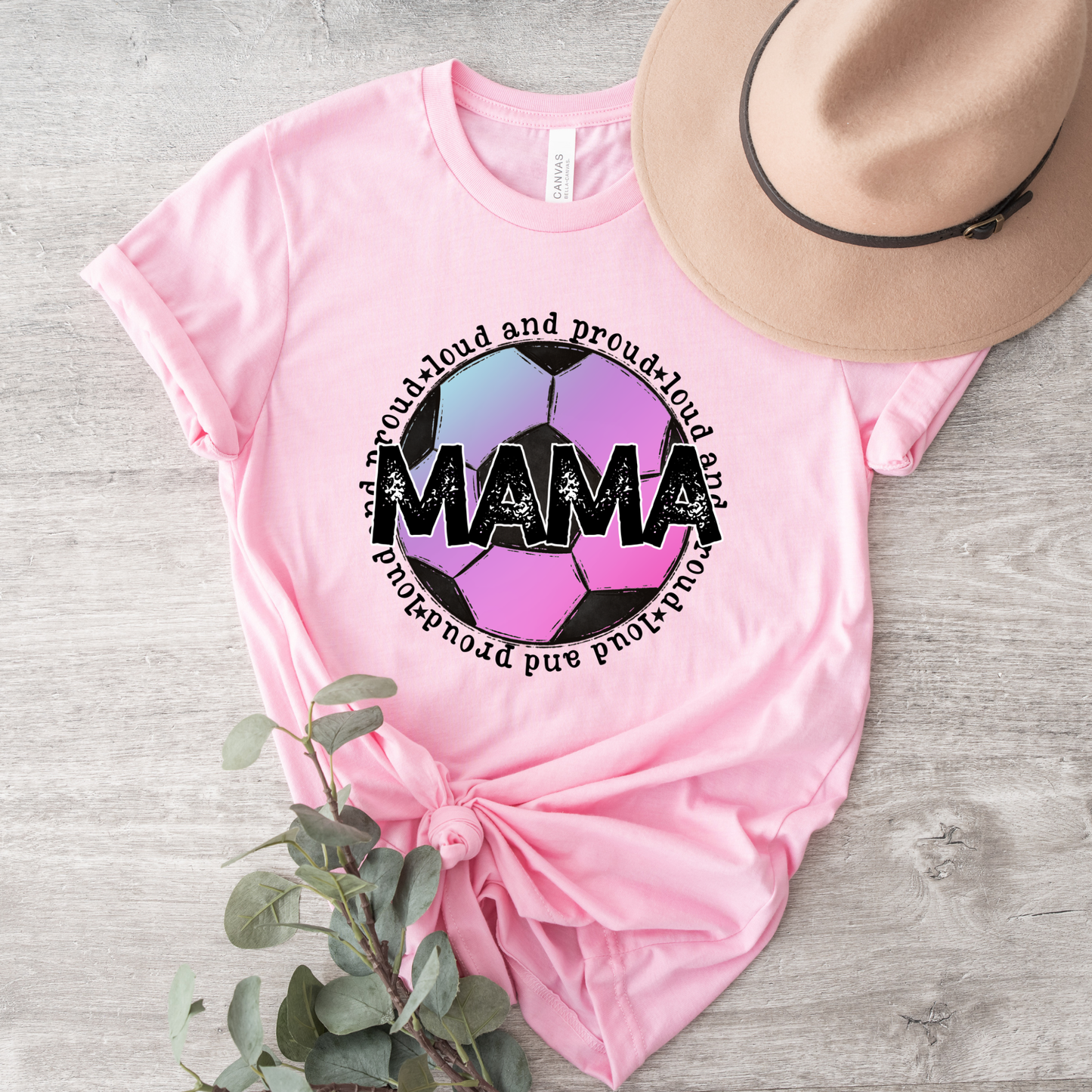A pink short-sleeve graphic T-shirt laid flat on a light wood surface, styled with a tan hat and greenery. The shirt has a large circular soccer-ball-style graphic with the word "MAMA" in black and pastel pink, purple, and blue sections, surrounded by curved text reading "proud and proud for MAMA.
