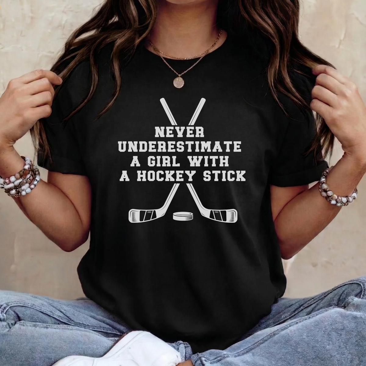 A woman wearing a black graphic t-shirt with white text that reads 'NEVER UNDERESTIMATE A GIRL WITH A HOCKEY STICK' above crossed hockey sticks and a puck. She is wearing light blue jeans and bracelets, sitting against a neutral beige wall.