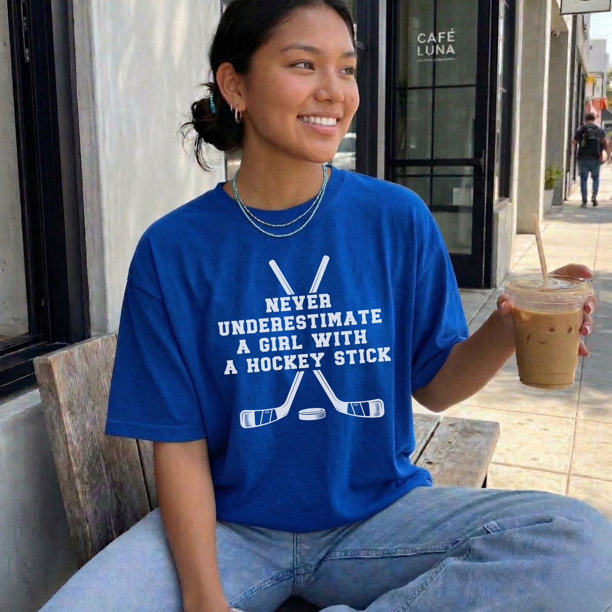 A woman sitting on a wooden bench at a cafe, wearing a bright blue graphic t-shirt with white text that reads 'NEVER UNDERESTIMATE A GIRL WITH A HOCKEY STICK' above crossed hockey sticks. She is holding an iced coffee.