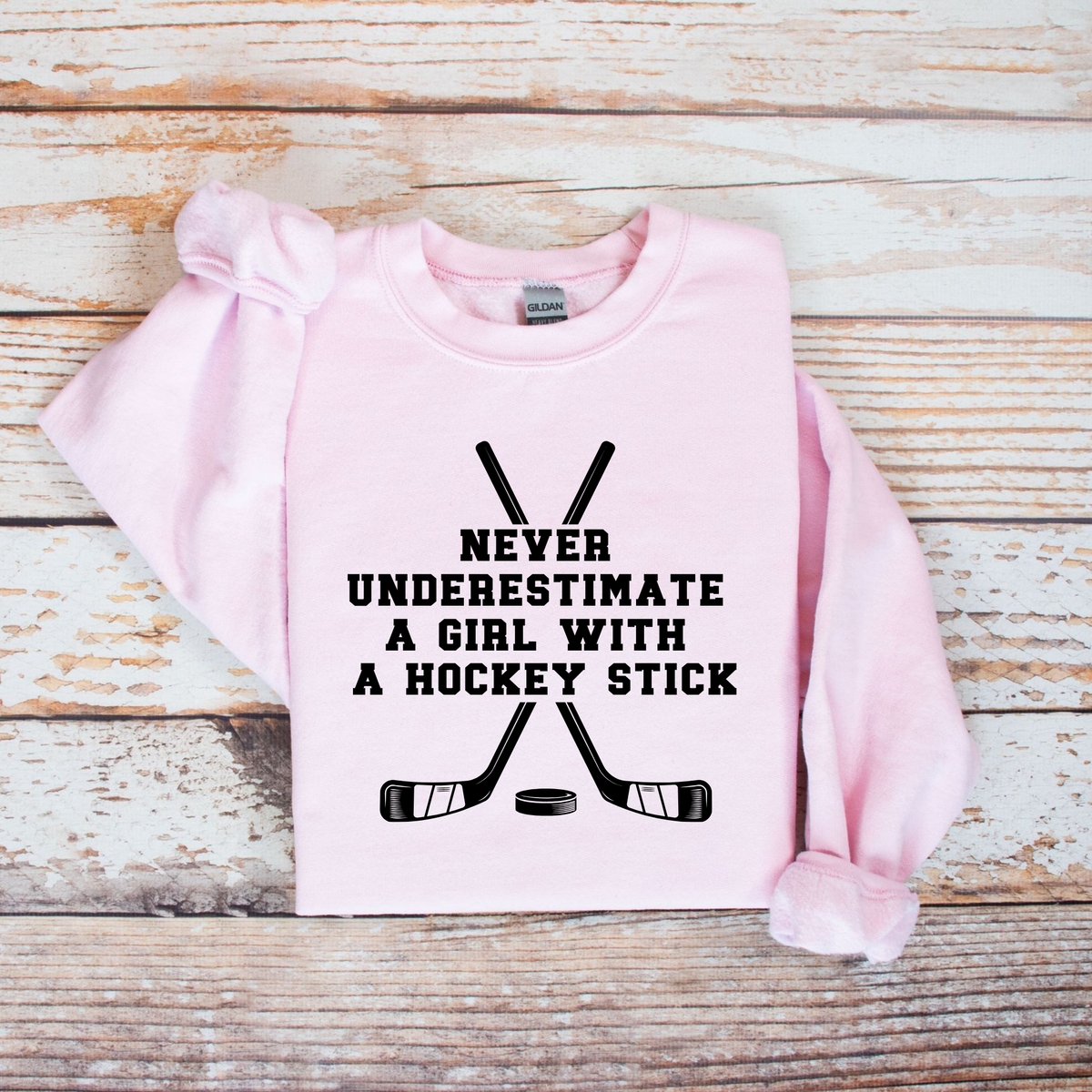 A light pink long-sleeve sweatshirt with rolled cuffs, featuring a black graphic of crossed hockey sticks and the text 'NEVER UNDERESTIMATE A GIRL WITH A HOCKEY STICK' on the front. The sweatshirt is laid flat on a rustic, whitewashed wooden plank background.