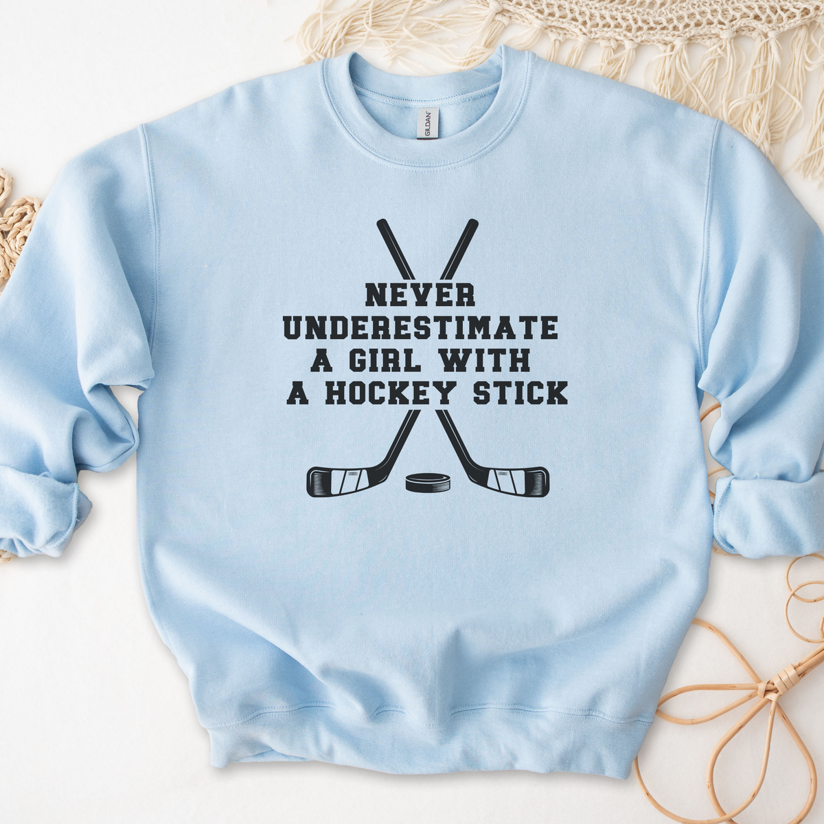 A light blue crewneck sweatshirt with a black graphic of crossed hockey sticks and the text 'NEVER UNDERESTIMATE A GIRL WITH A HOCKEY STICK' printed on the chest, displayed flat on a white surface with neutral decorative props.