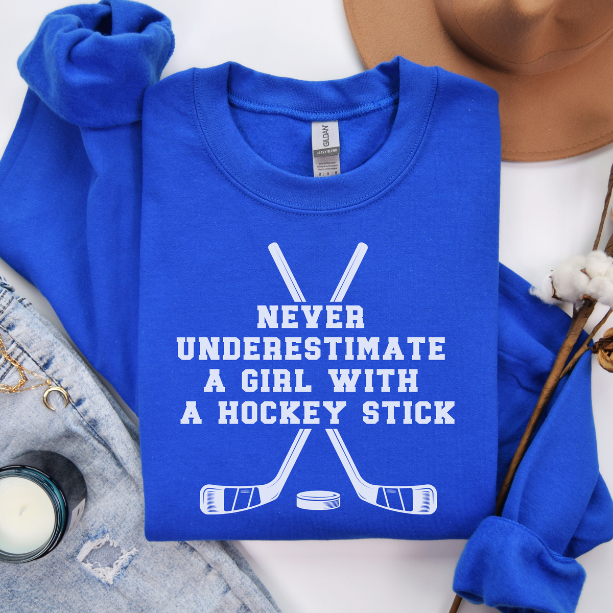 A folded royal blue sweatshirt with a white hockey graphic and the text 'NEVER UNDERESTIMATE A GIRL WITH A HOCKEY STICK' on the front, styled in a flat lay with a matching sweatshirt, denim, a hat, and dried flowers.