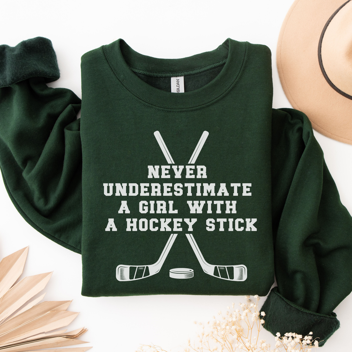 A dark green long-sleeve sweatshirt with a white graphic text reading 'NEVER UNDERESTIMATE A GIRL WITH A HOCKEY STICK' above crossed hockey sticks and a puck, styled in a flat lay with a tan hat, dried flowers, and wooden leaves on a white background.