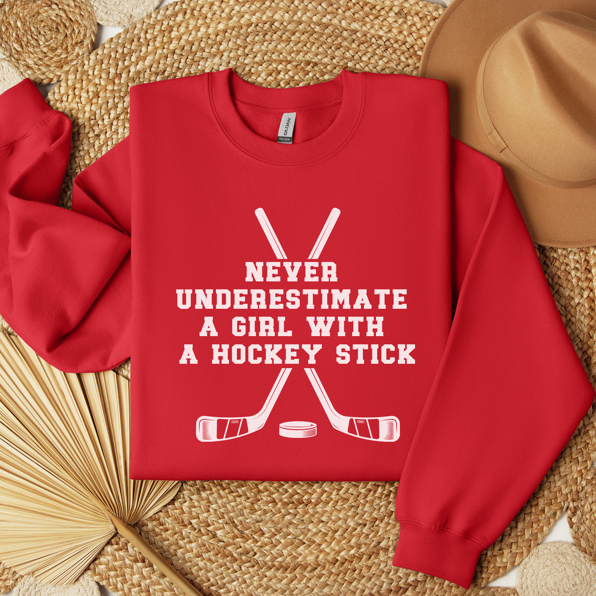 A red crewneck sweatshirt with a white graphic print reading 'NEVER UNDERESTIMATE A GIRL WITH A HOCKEY STICK' around crossed hockey sticks and a puck. The sweatshirt is displayed in a flat-lay arrangement on a woven straw mat with a tan hat and dried palm leaves.
