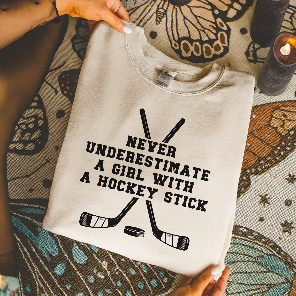 A folded light beige crewneck sweatshirt with a black graphic of crossed hockey sticks and the text 'NEVER UNDERESTIMATE A GIRL WITH A HOCKEY STICK' is being held by hands over a patterned rug.