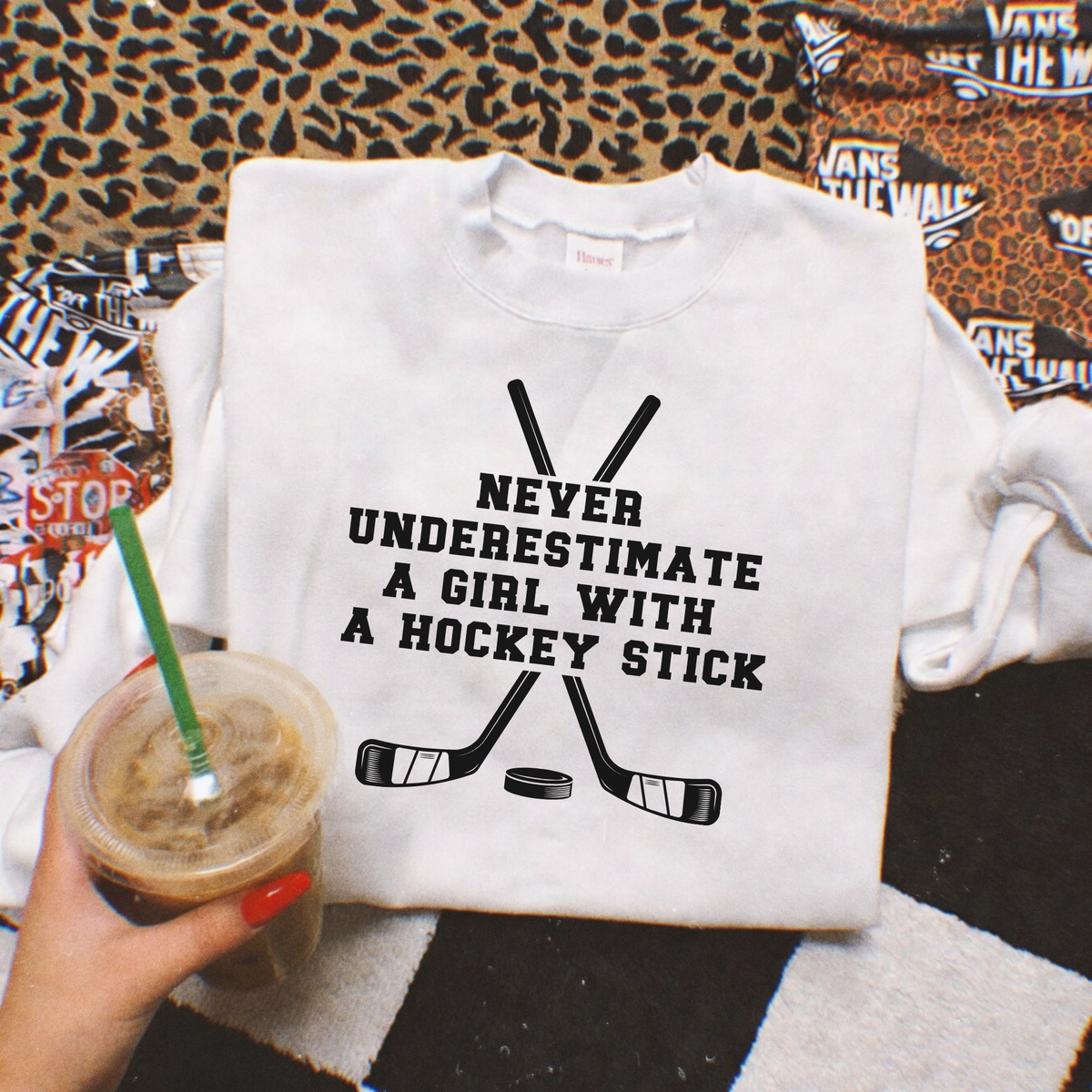 A white sweatshirt with a black graphic of crossed hockey sticks and the text 'NEVER UNDERESTIMATE A GIRL WITH A HOCKEY STICK' is laid flat on a patterned surface. A hand holding an iced coffee is visible in the lower left corner.