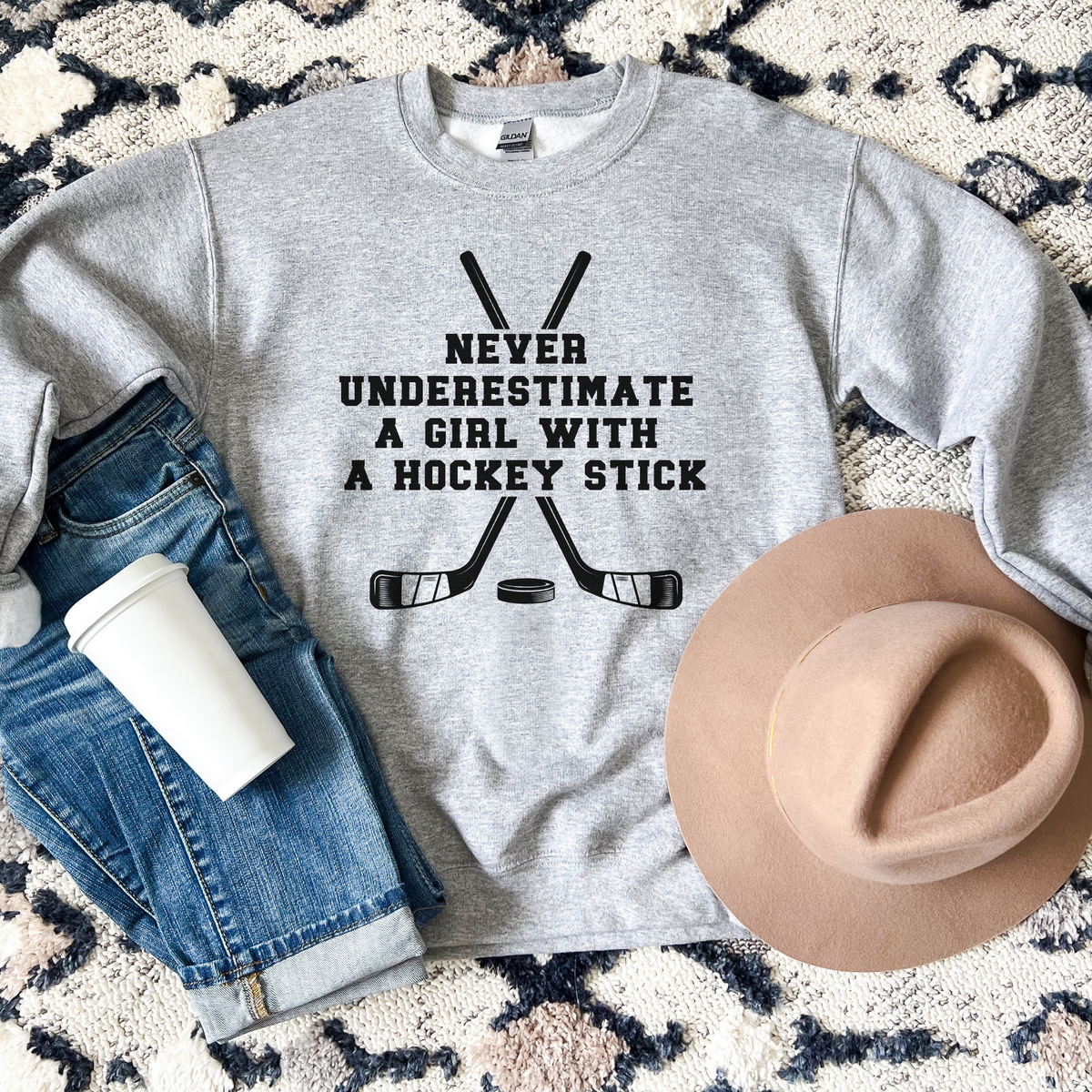 A heather gray crewneck sweatshirt with a black graphic reading 'NEVER UNDERESTIMATE A GIRL WITH A HOCKEY STICK' above crossed hockey sticks and a puck. The sweatshirt is styled in a flat lay with blue jeans, a white coffee cup, and a tan hat on a patterned rug.
