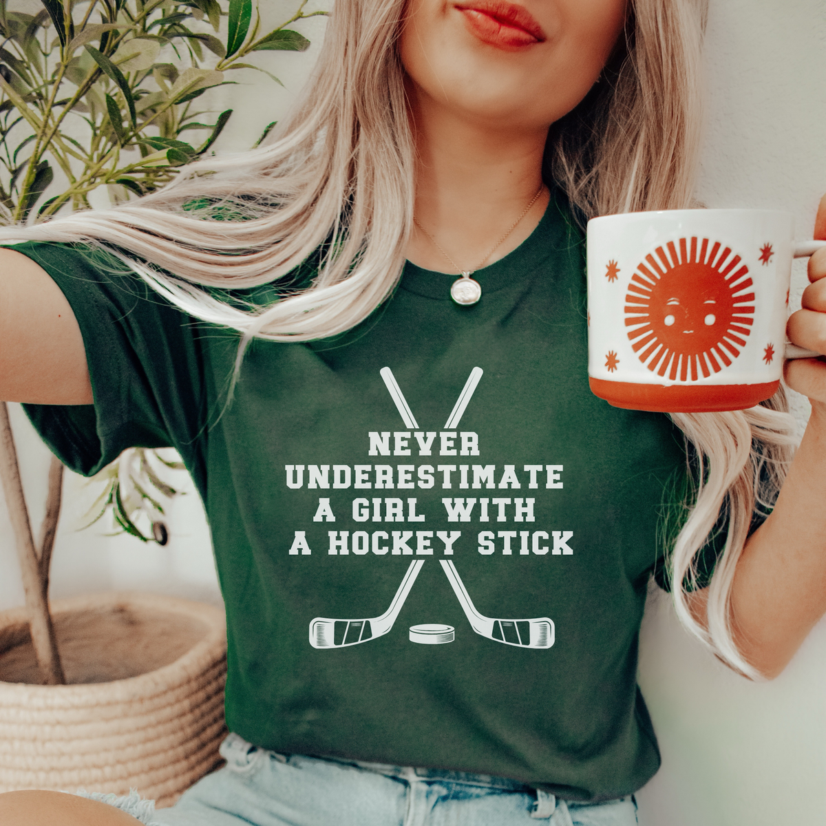 A woman wearing a dark green graphic t-shirt with the text 'NEVER UNDERESTIMATE A GIRL WITH A HOCKEY STICK' and crossed hockey sticks printed on the front. She is holding a white ceramic mug with a red sun graphic, with a potted plant in the background.