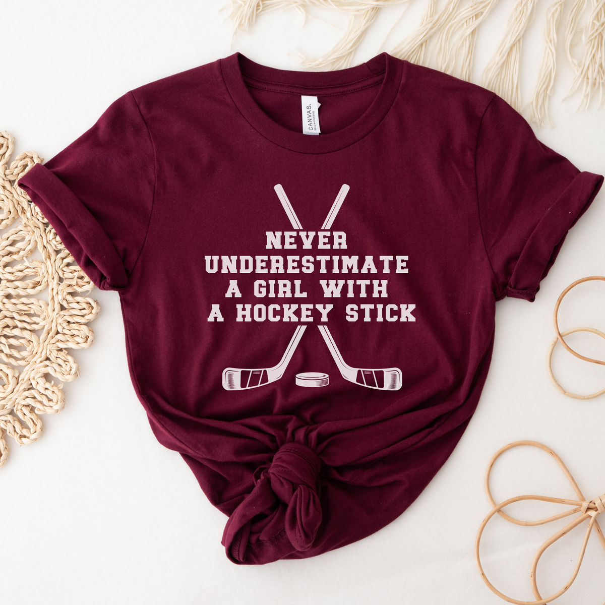 A maroon short-sleeve t-shirt with a white graphic print that reads 'NEVER UNDERESTIMATE A GIRL WITH A HOCKEY STICK' above crossed hockey sticks and a puck. The shirt is styled in a flat lay with a knot at the bottom, surrounded by neutral decorative props.