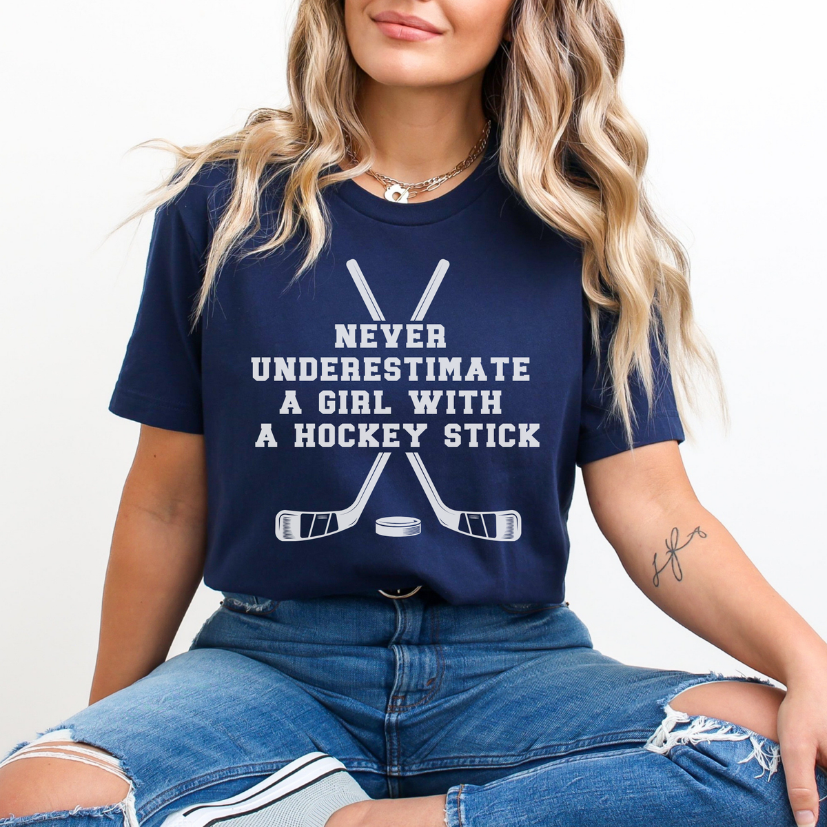 A woman wearing a navy blue graphic t-shirt with white text reading 'NEVER UNDERESTIMATE A GIRL WITH A HOCKEY STICK' and crossed hockey sticks, paired with ripped blue jeans against a plain white background.