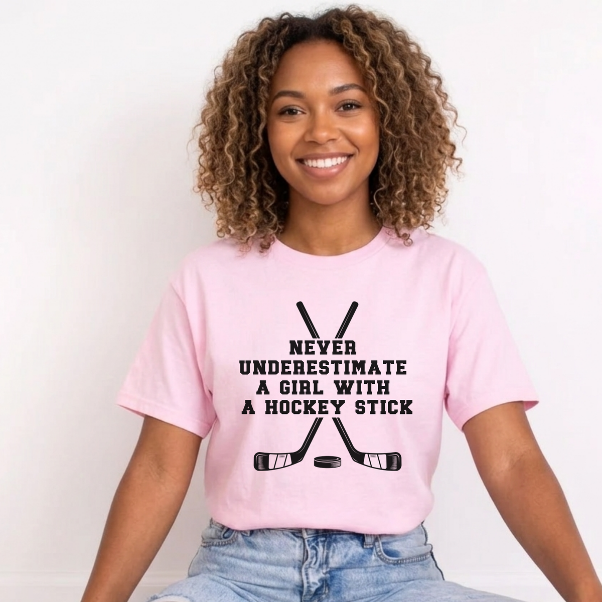 A smiling woman with curly brown hair wearing a light pink graphic t-shirt and light wash blue jeans, sitting against a plain white background.