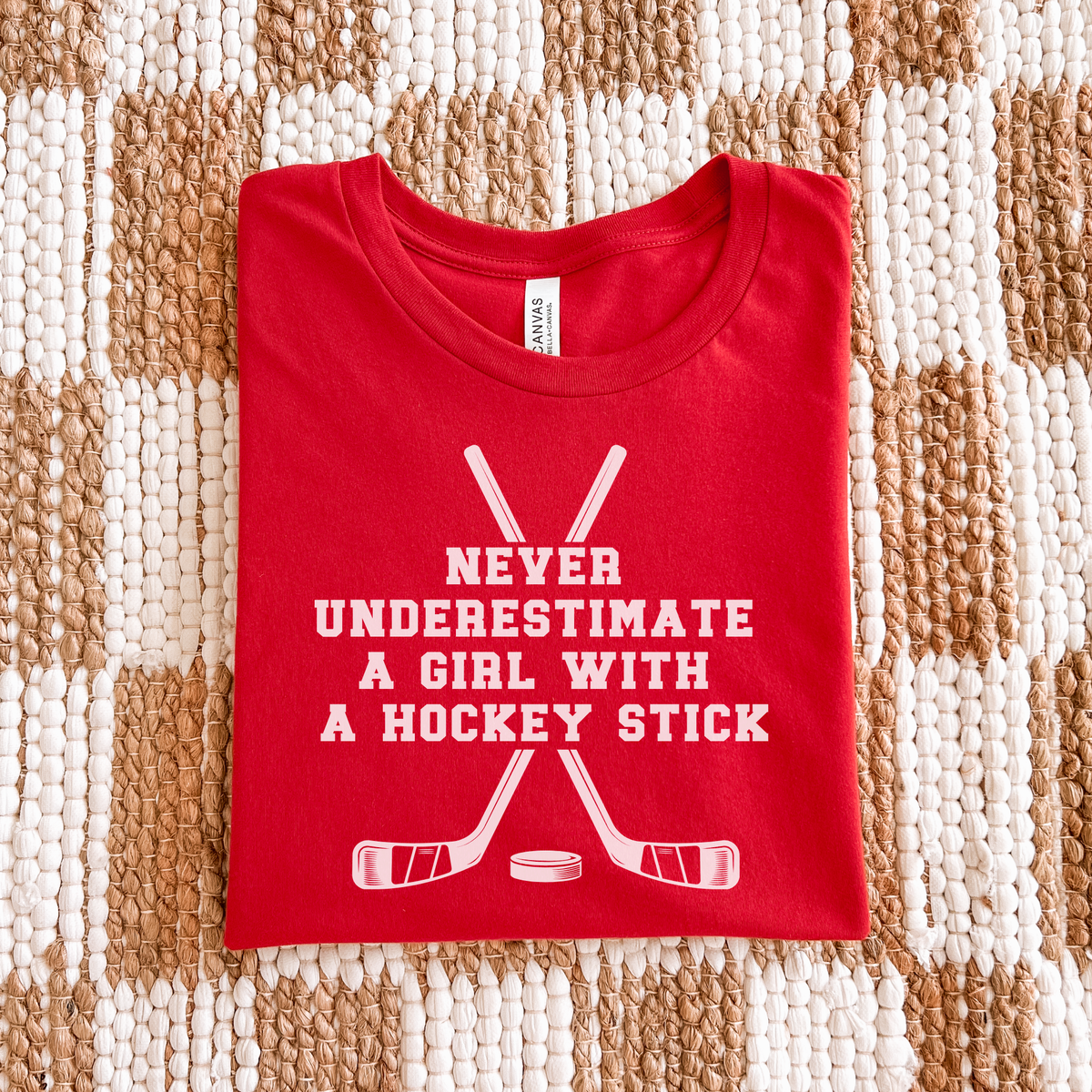 A folded red t-shirt with a white graphic print that reads 'NEVER UNDERESTIMATE A GIRL WITH A HOCKEY STICK' above crossed hockey sticks and a puck, laid flat on a beige and white woven rug.