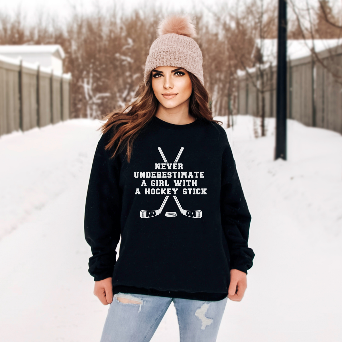 A woman standing outdoors in the snow wearing a black crewneck sweatshirt with a white graphic that reads 'NEVER UNDERESTIMATE A GIRL WITH A HOCKEY STICK' above crossed hockey sticks, paired with light blue ripped jeans and a beige knit pom-pom beanie.