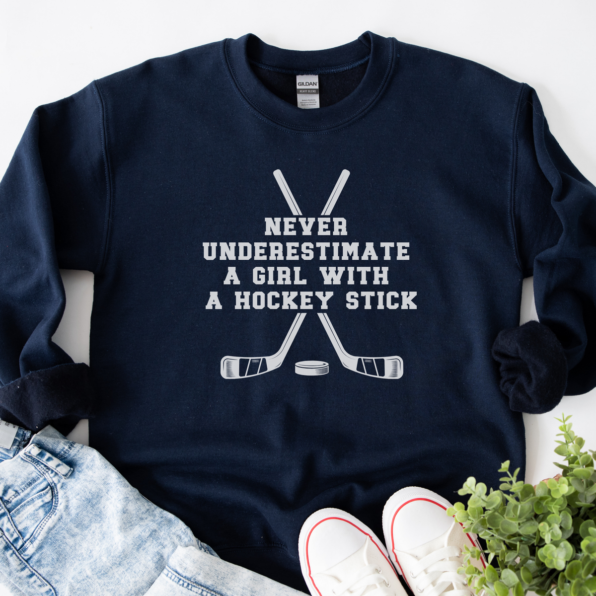 A navy blue crewneck sweatshirt with a white graphic print that reads 'NEVER UNDERESTIMATE A GIRL WITH A HOCKEY STICK' above crossed hockey sticks and a puck. The sweatshirt is styled in a flat lay with light-wash denim jeans, white sneakers, and a small green plant on a white background.