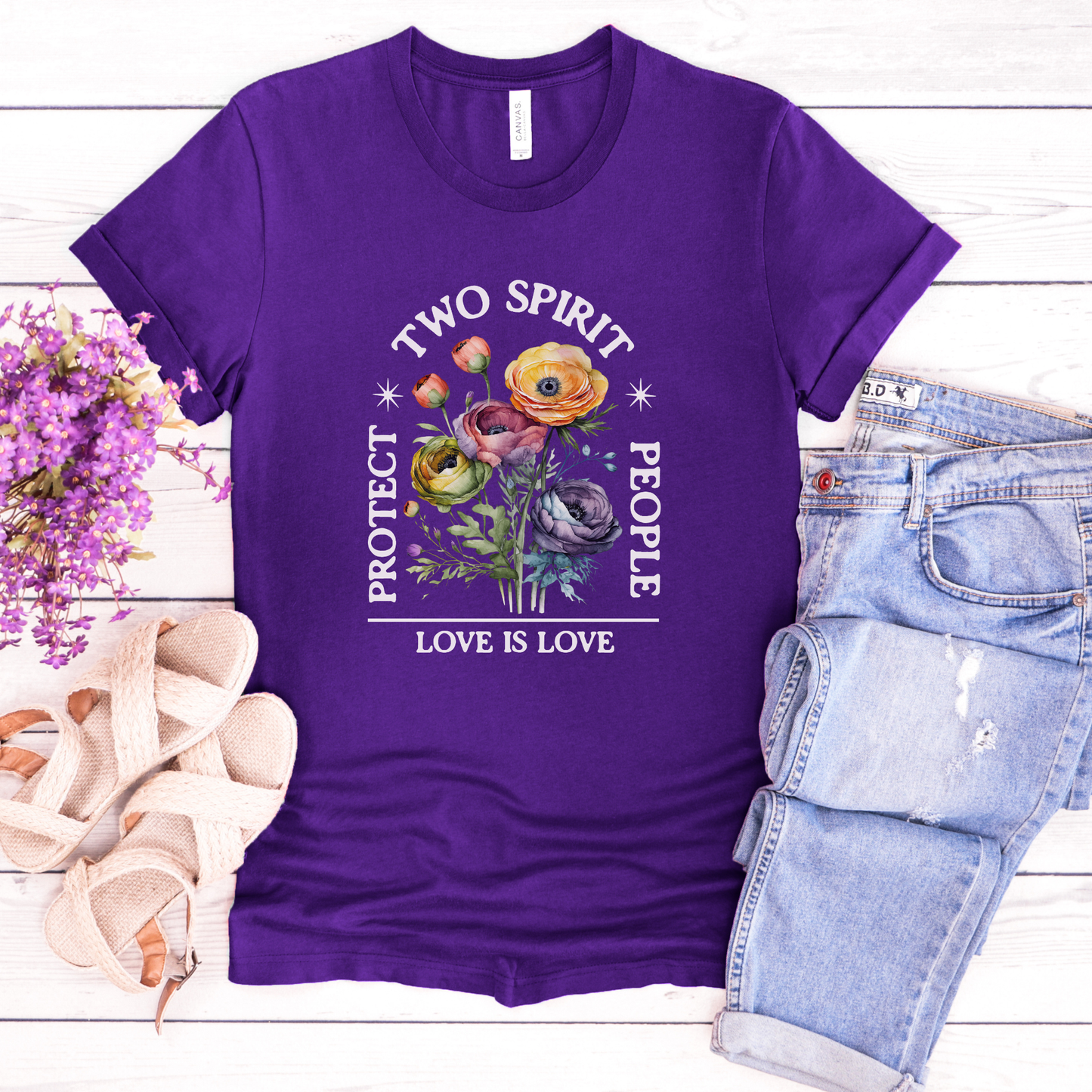 Purple t-shirt with 'Two Spirit People, Love is Love' design on a wooden surface with flowers and jeans. protect two spirit people shirt, love is love two spirit tee, indigenous lgbtq shirt, two spirit pride t-shirt, indigenous pride apparel, lgbtq indigenous tee, two spirit advocacy shirt, love is love unisex tee, 2s pride shirt, indigenous lgbtq clothing, two spirit protection tee, queer indigenous apparel