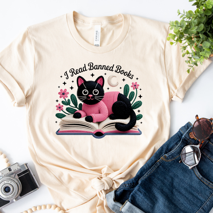Beige t-shirt with a black cat reading a book graphic and 'I Read Banned Books' text, placed on a white surface with jeans and a camera.